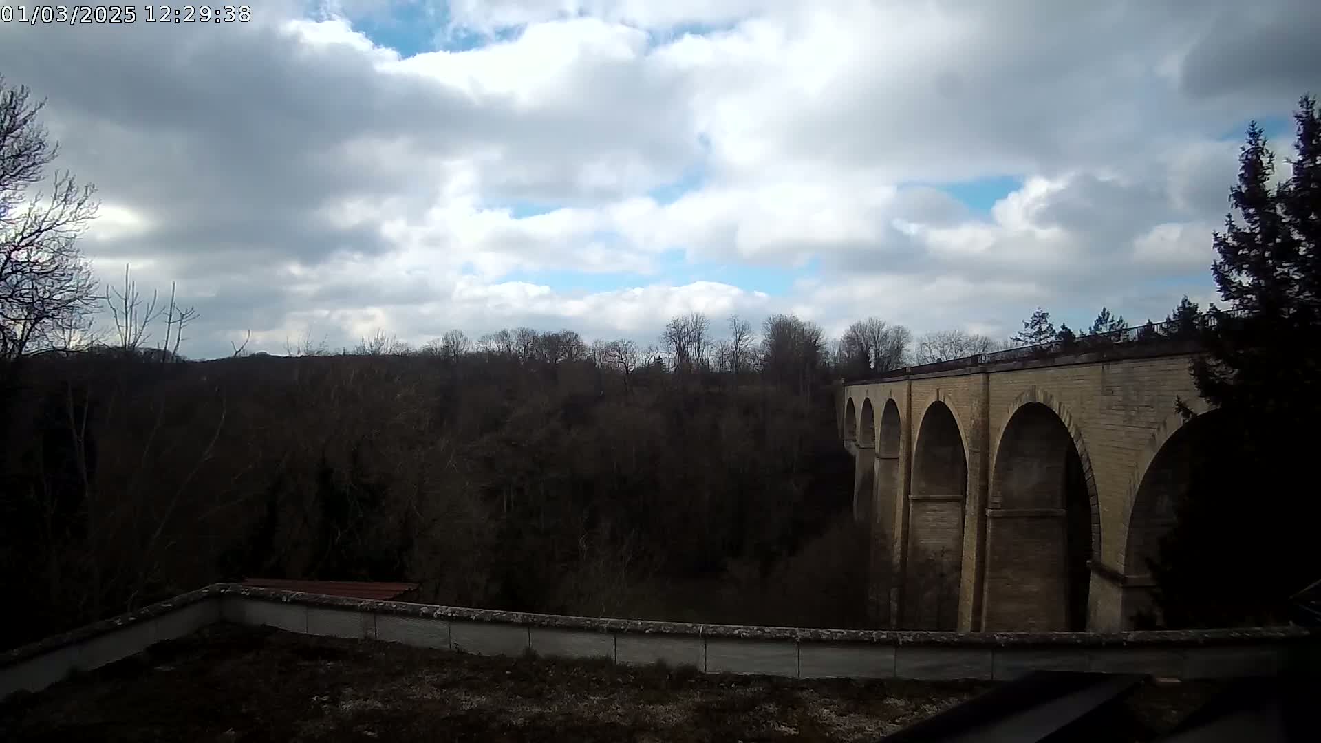A stone viaduct with multiple arches spans a wooded valley under a partly cloudy sky.