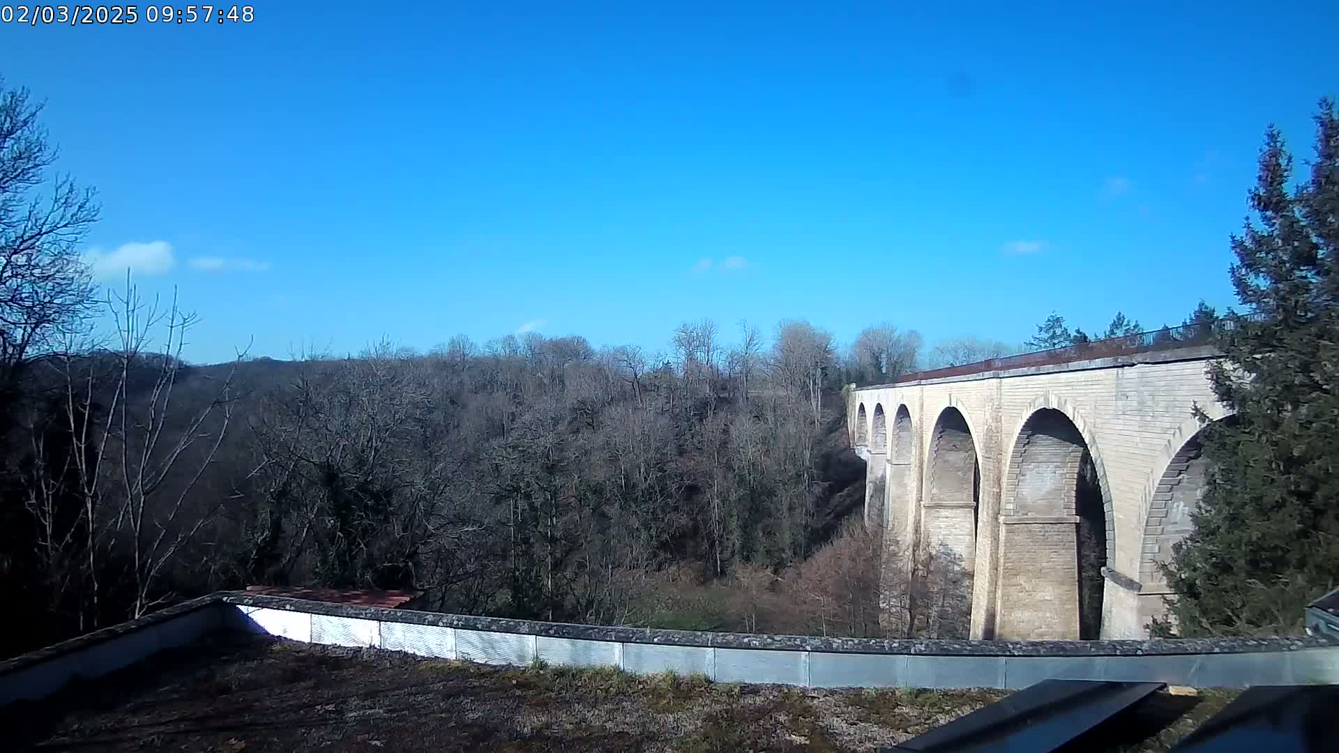 A long stone viaduct with multiple arches stretches across a valley of leafless trees under a clear blue sky.