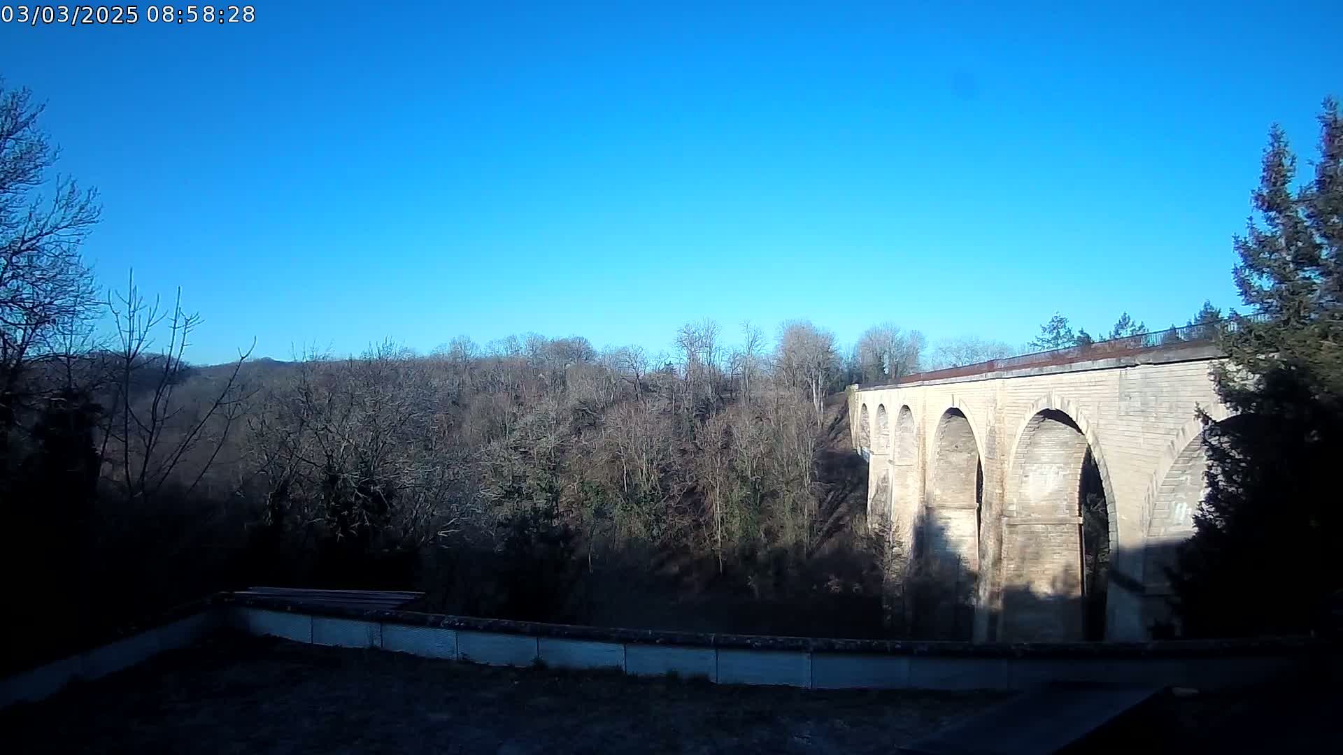A stone viaduct with multiple arches stands against a clear blue sky, overlooking a winter forest.