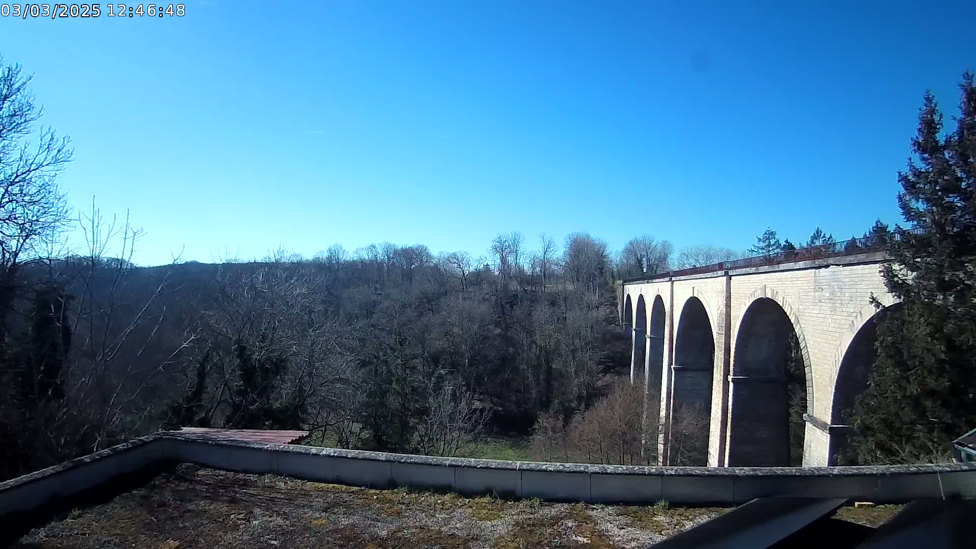 A stone viaduct with multiple arches is visible beyond a low wall and a flat, vegetated rooftop, all under a clear blue sky.