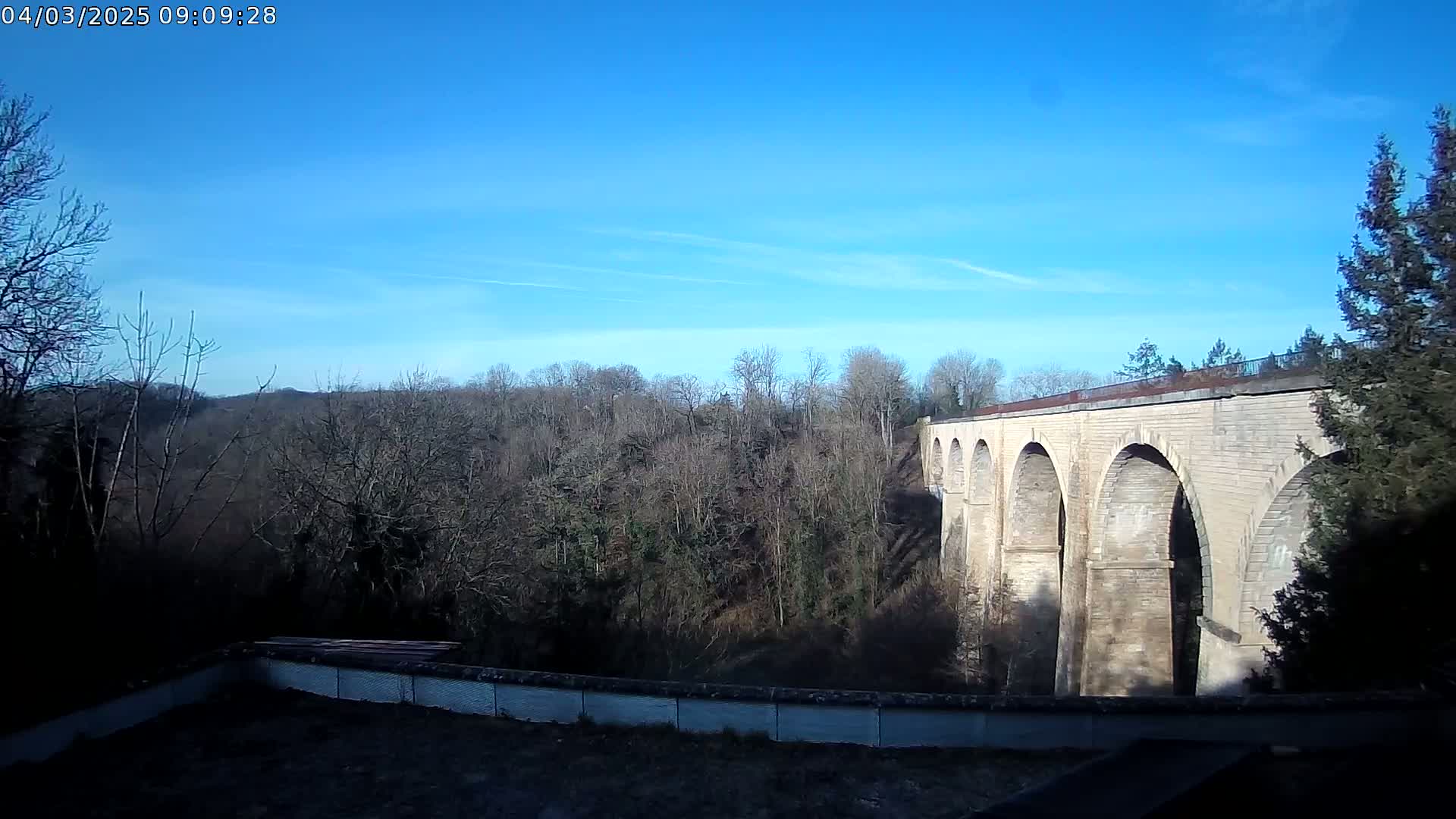 A stone viaduct with multiple arches stretches across a valley of leafless trees under a clear blue sky.