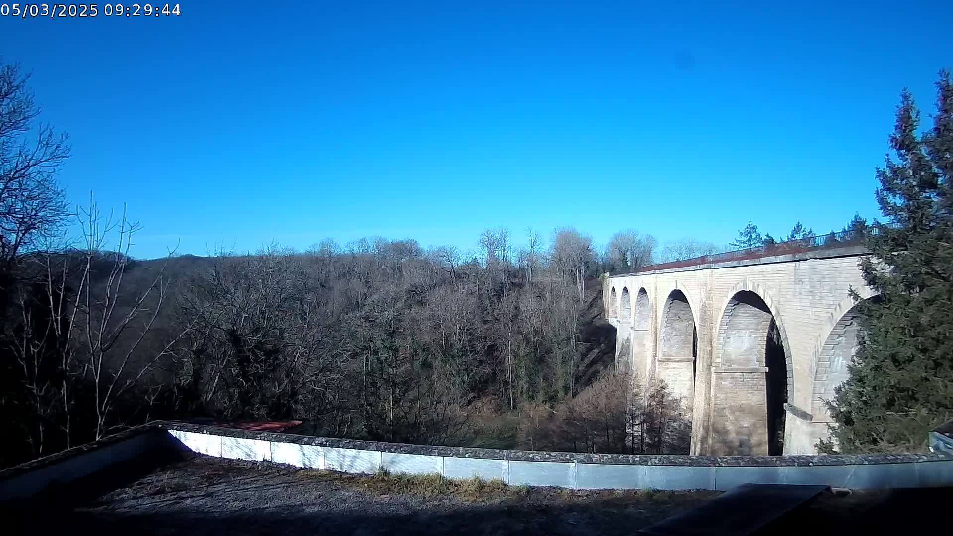 A stone viaduct with multiple arches is visible beyond a low wall and a background of leafless trees under a clear blue sky.