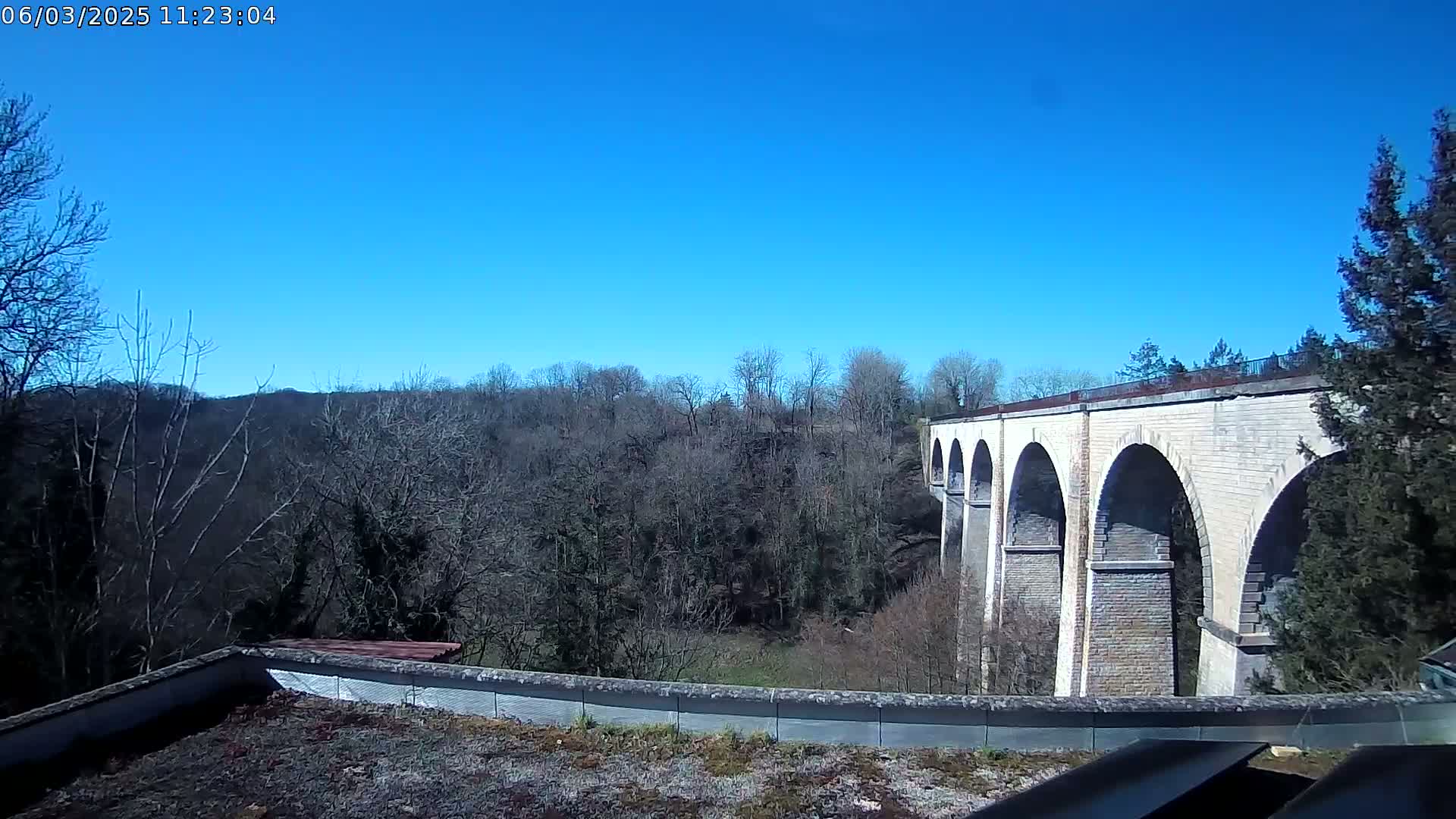 A stone viaduct with multiple arches spans a valley of leafless trees under a clear blue sky.