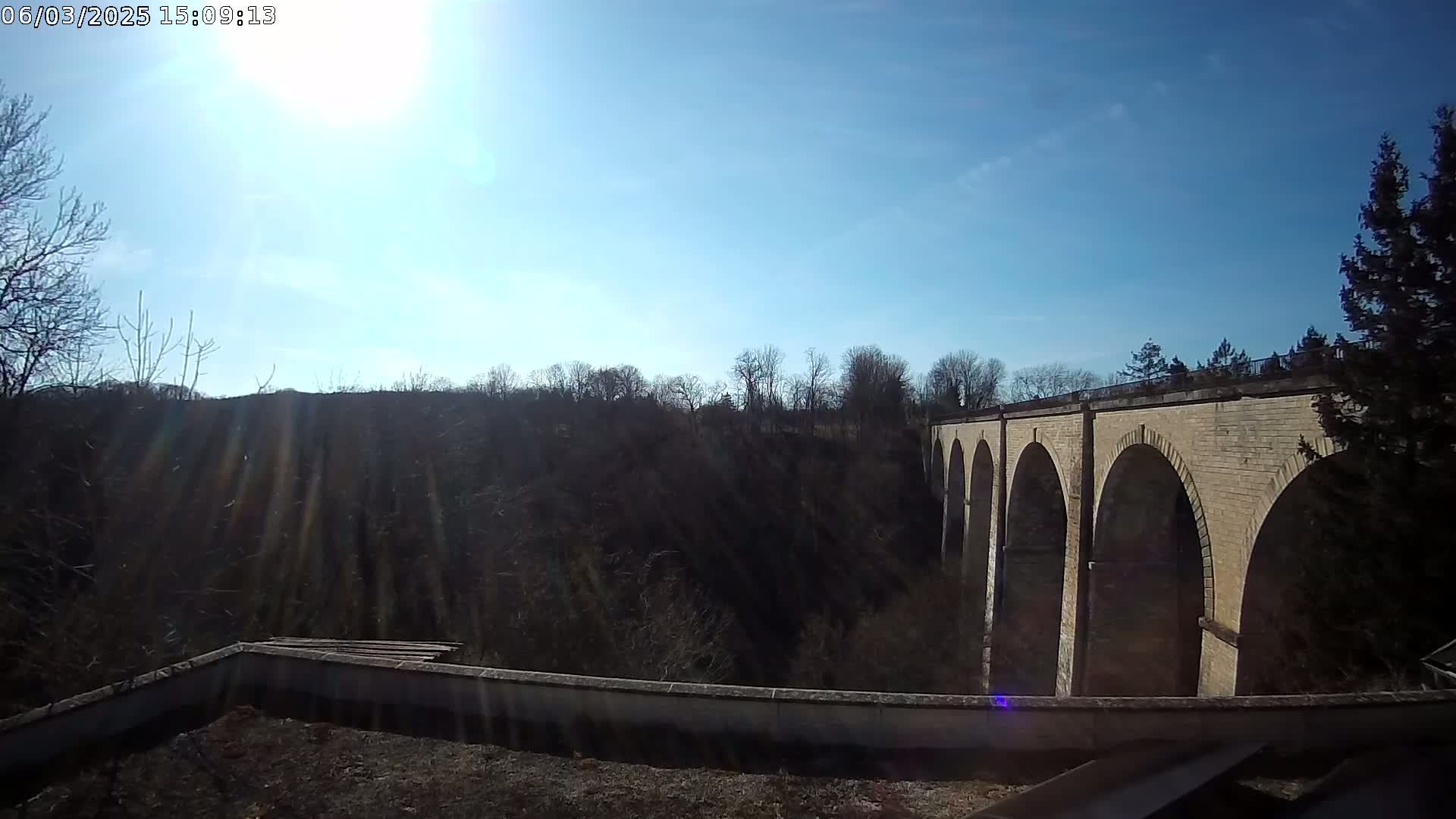 A stone bridge with multiple arches extends from a hilltop into a valley under a bright, sunny sky.