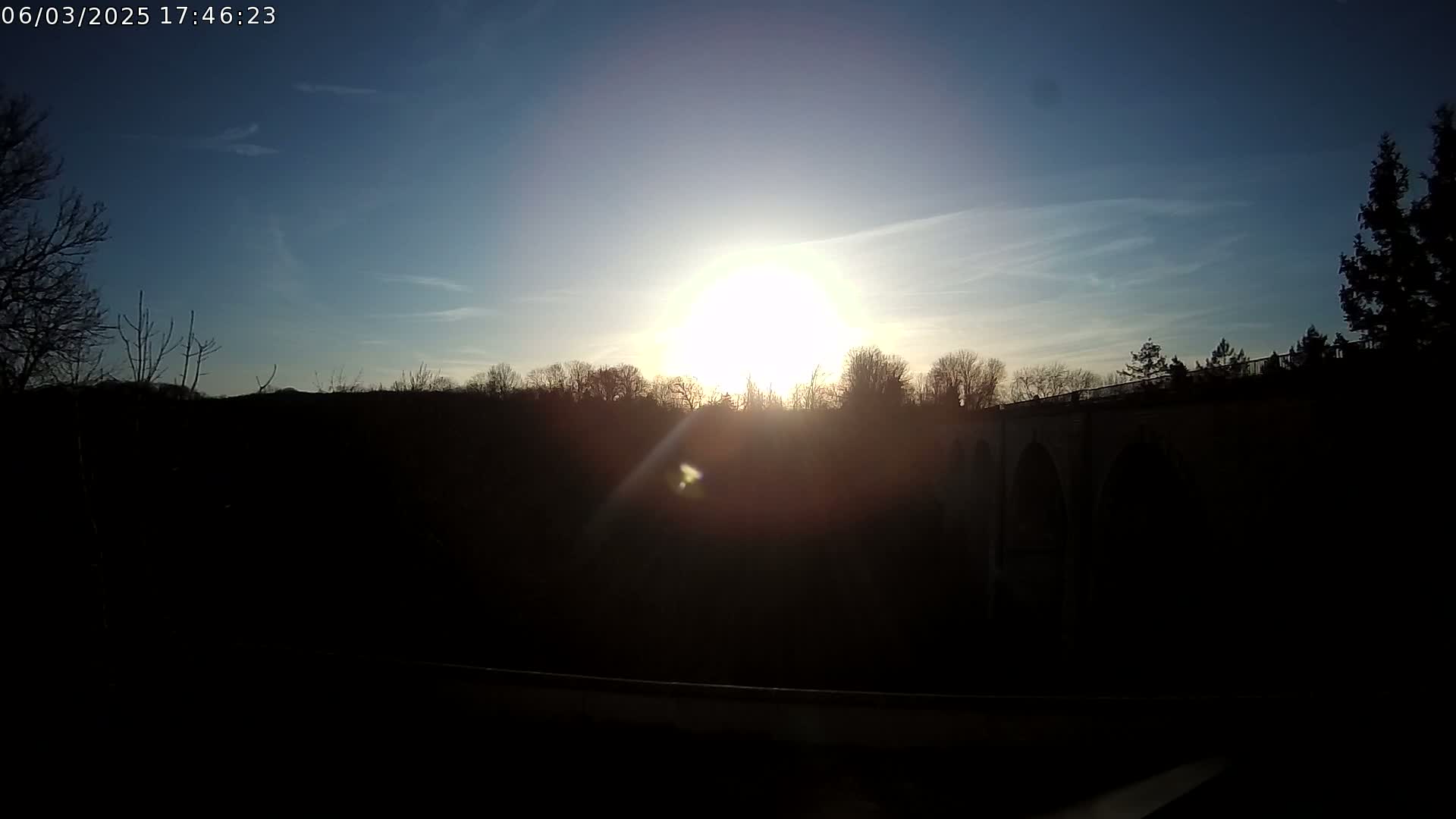 A bright sun shines over a dark silhouette of trees and a stone bridge under a mostly clear blue sky.