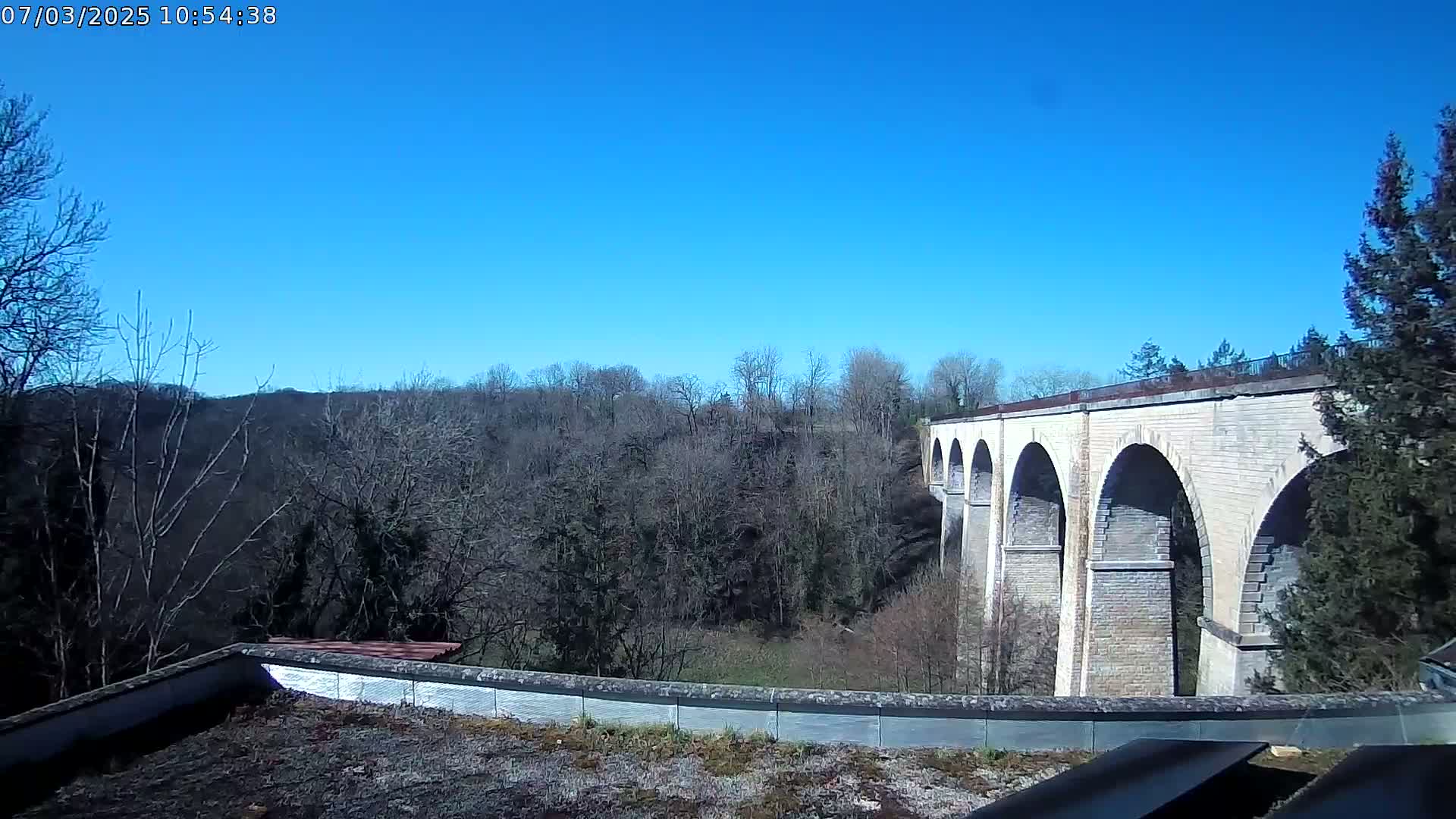 A stone viaduct with multiple arches spans a wooded valley under a clear blue sky.