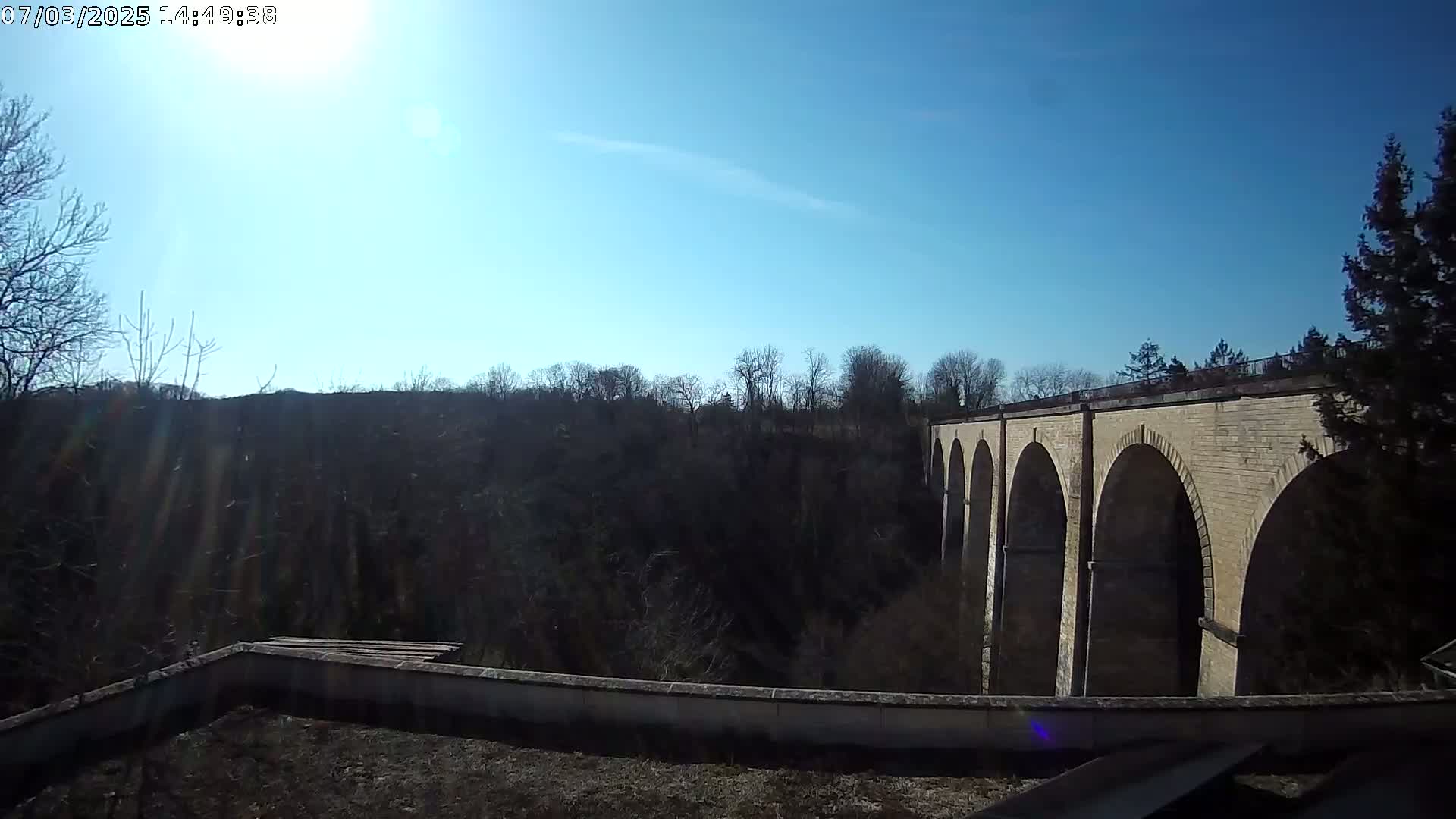 A stone viaduct with multiple arches is visible beyond a low wall, under a bright, sunny sky.
