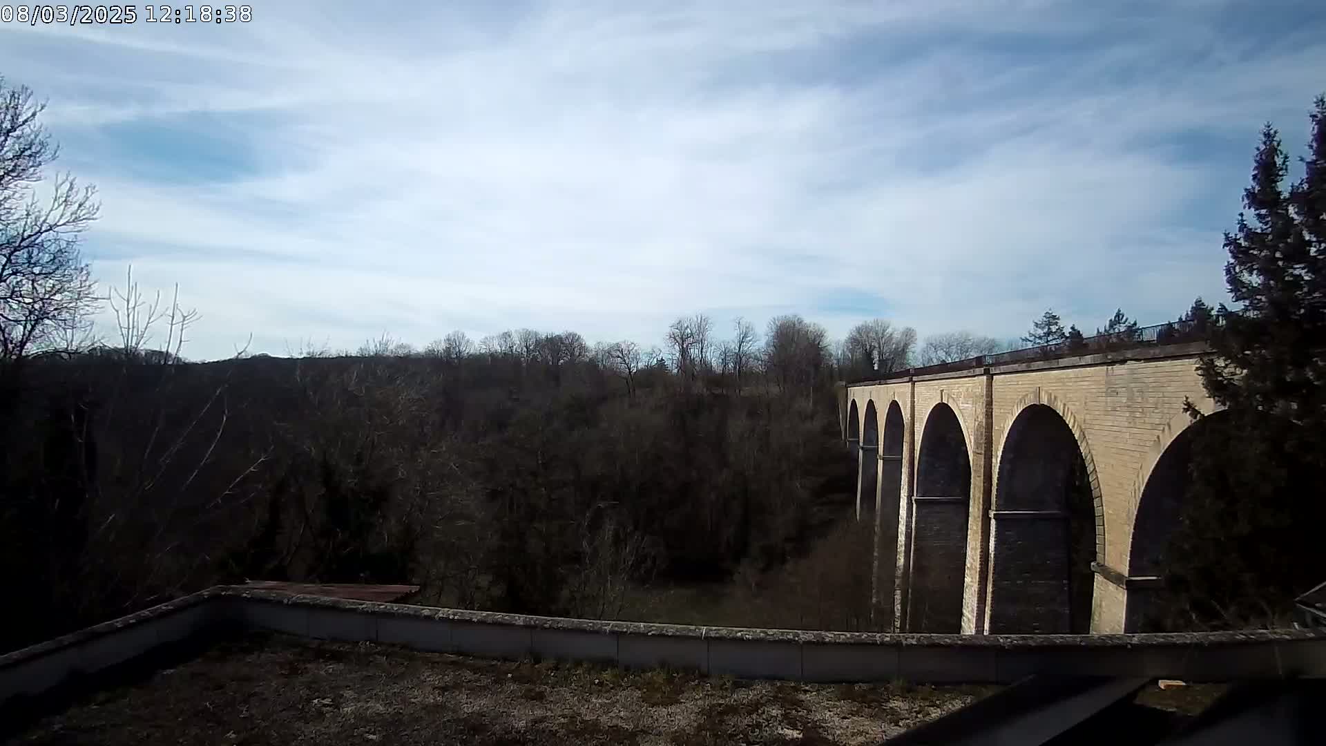 A stone viaduct with multiple arches stretches across a wooded valley under a partly cloudy sky.