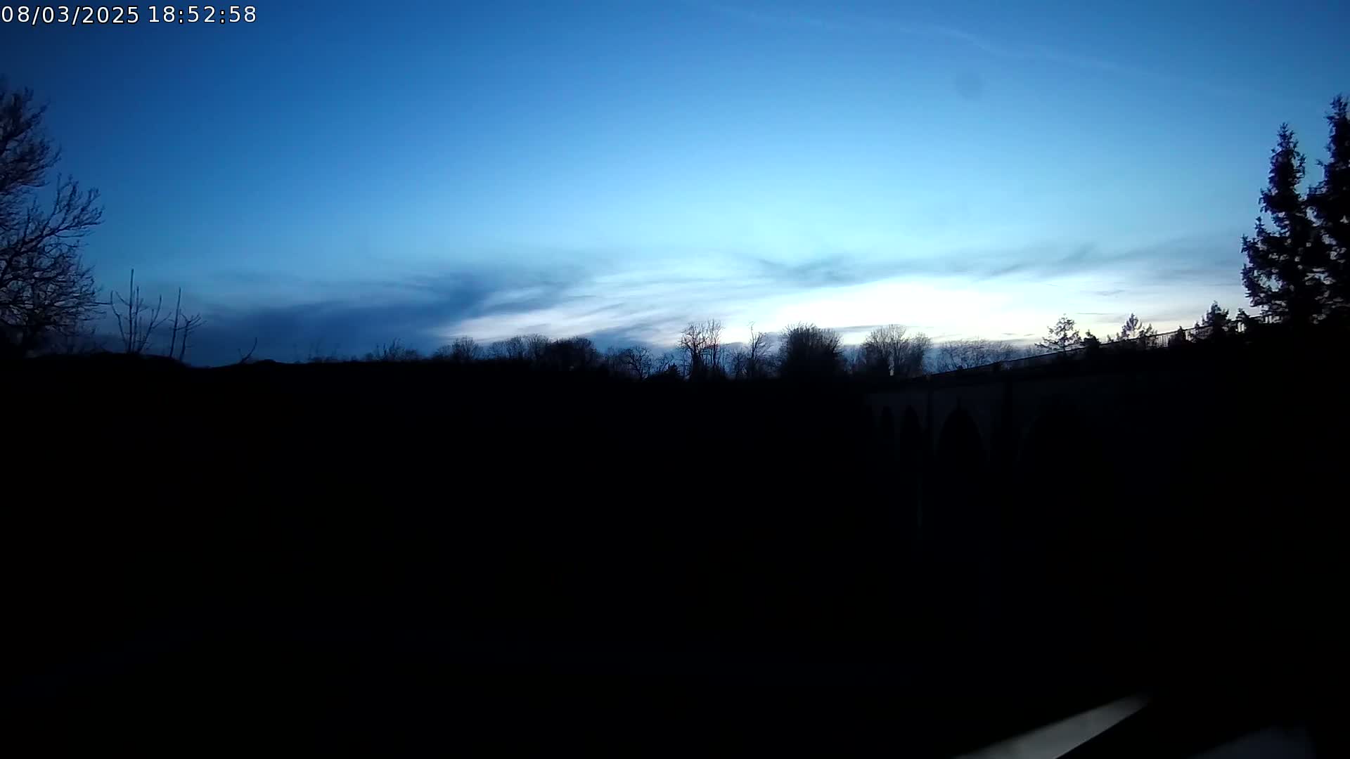 A twilight sky with thin clouds over a dark, silhouetted landscape featuring trees and a structure that may be a bridge or viaduct.