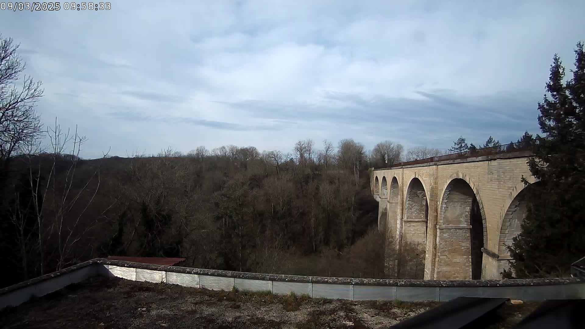 A stone viaduct stretches across a valley of leafless trees under a mostly cloudy sky.