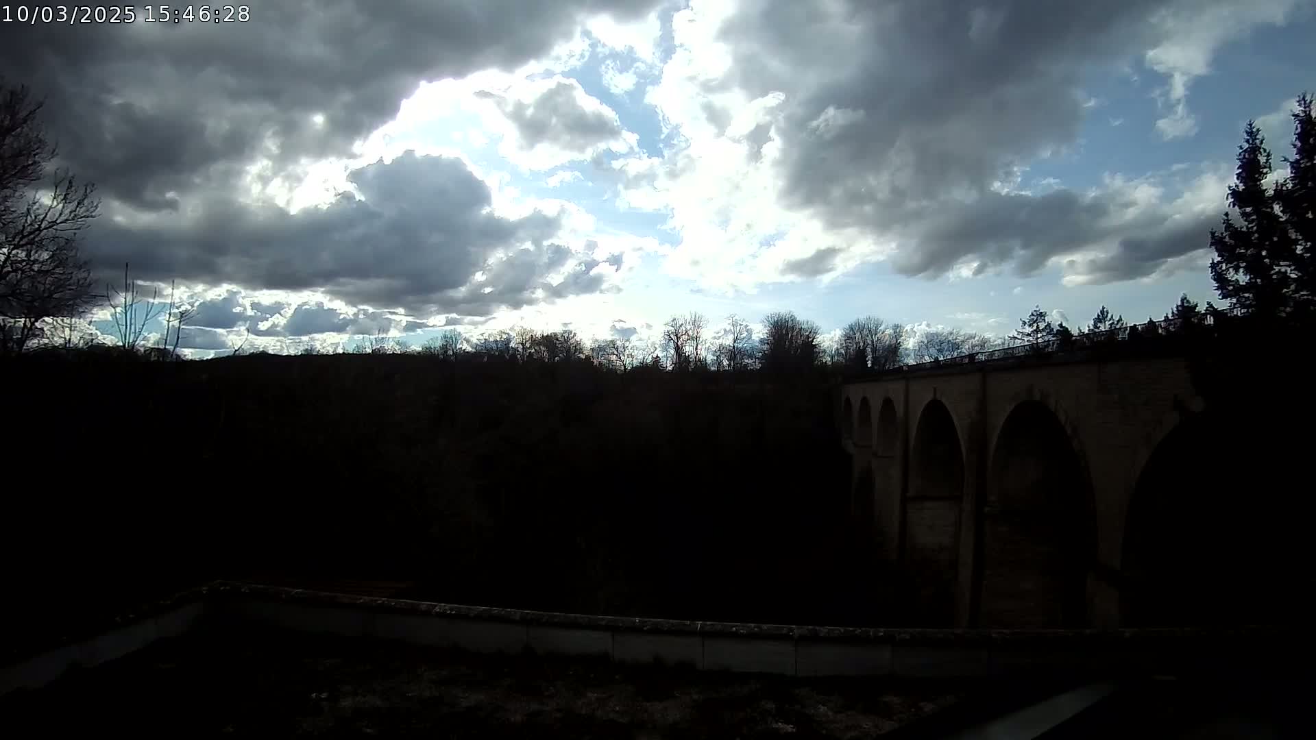 A stone bridge with multiple arches is partially visible behind a dark line of trees under a partly sunny sky with puffy clouds.