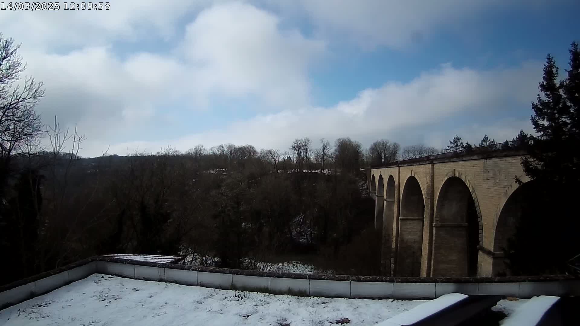 A snow-covered rooftop overlooks a stone viaduct, situated amidst a wintery landscape under a partly cloudy sky.