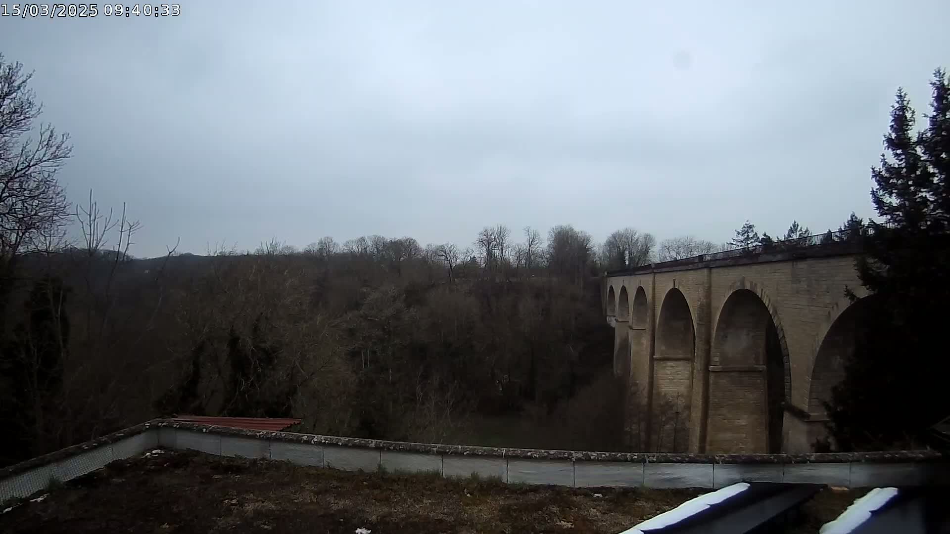 A stone viaduct with multiple arches is visible beyond a low wall and a line of bare winter trees under an overcast sky.