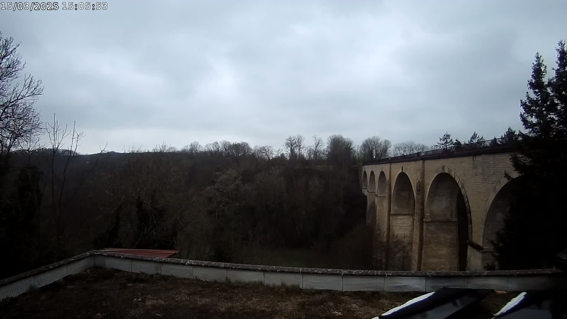 A stone viaduct with multiple arches is visible beyond a low wall and a wooded area under a cloudy sky.