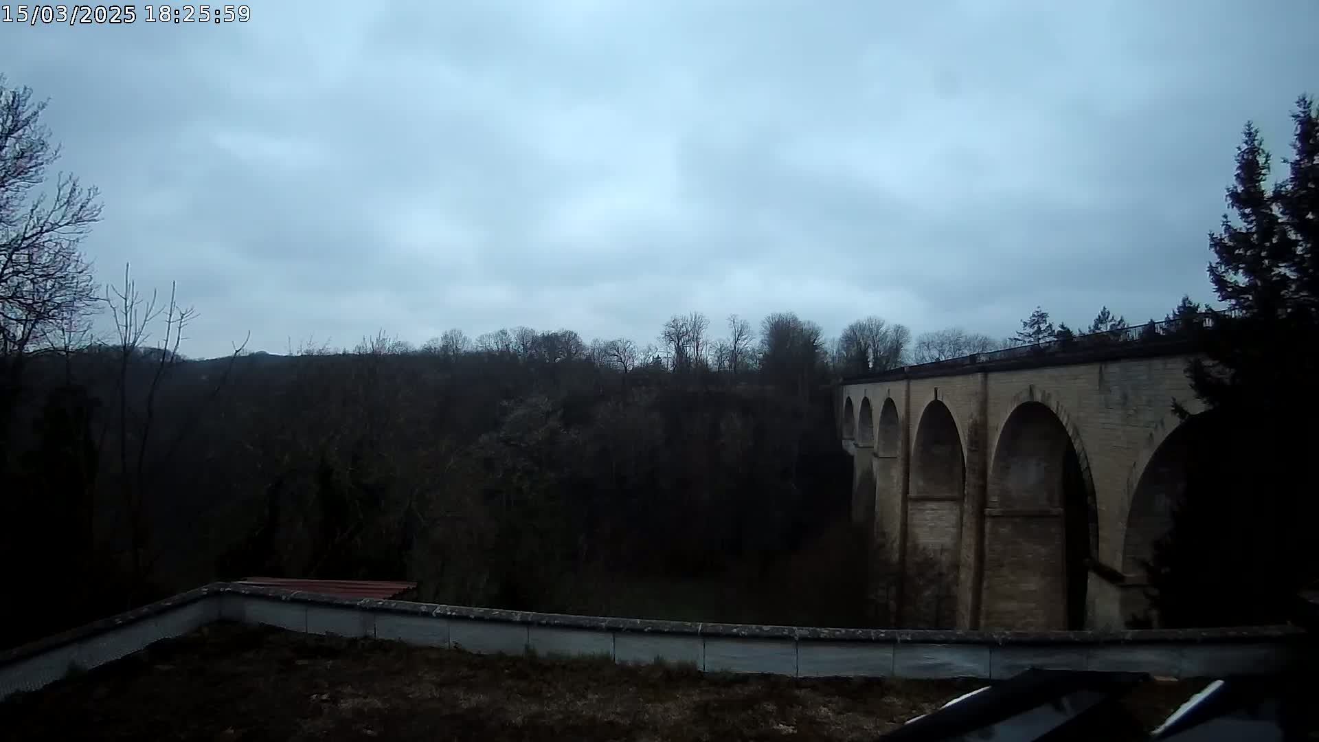 A stone viaduct with multiple arches is visible beyond a low wall and a sparsely vegetated area under an overcast sky.