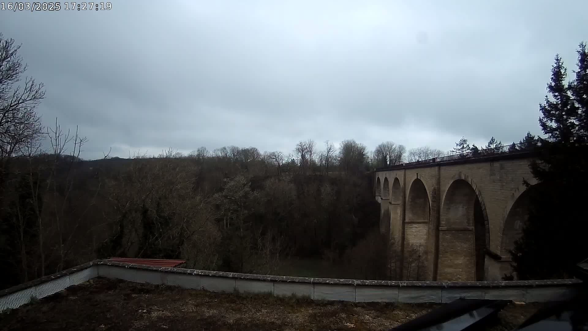 A stone viaduct with multiple arches stretches across a valley filled with leafless trees under an overcast sky.