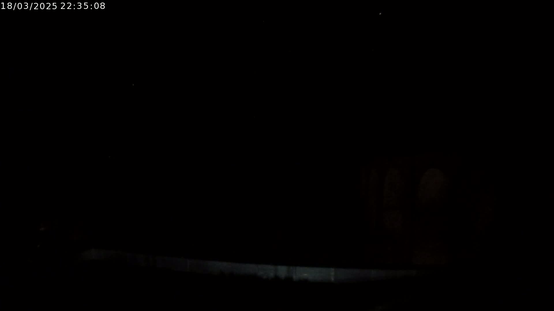 A dark, nighttime scene shows a low-light view of a blurry, indistinct object near a low wall or barrier.
