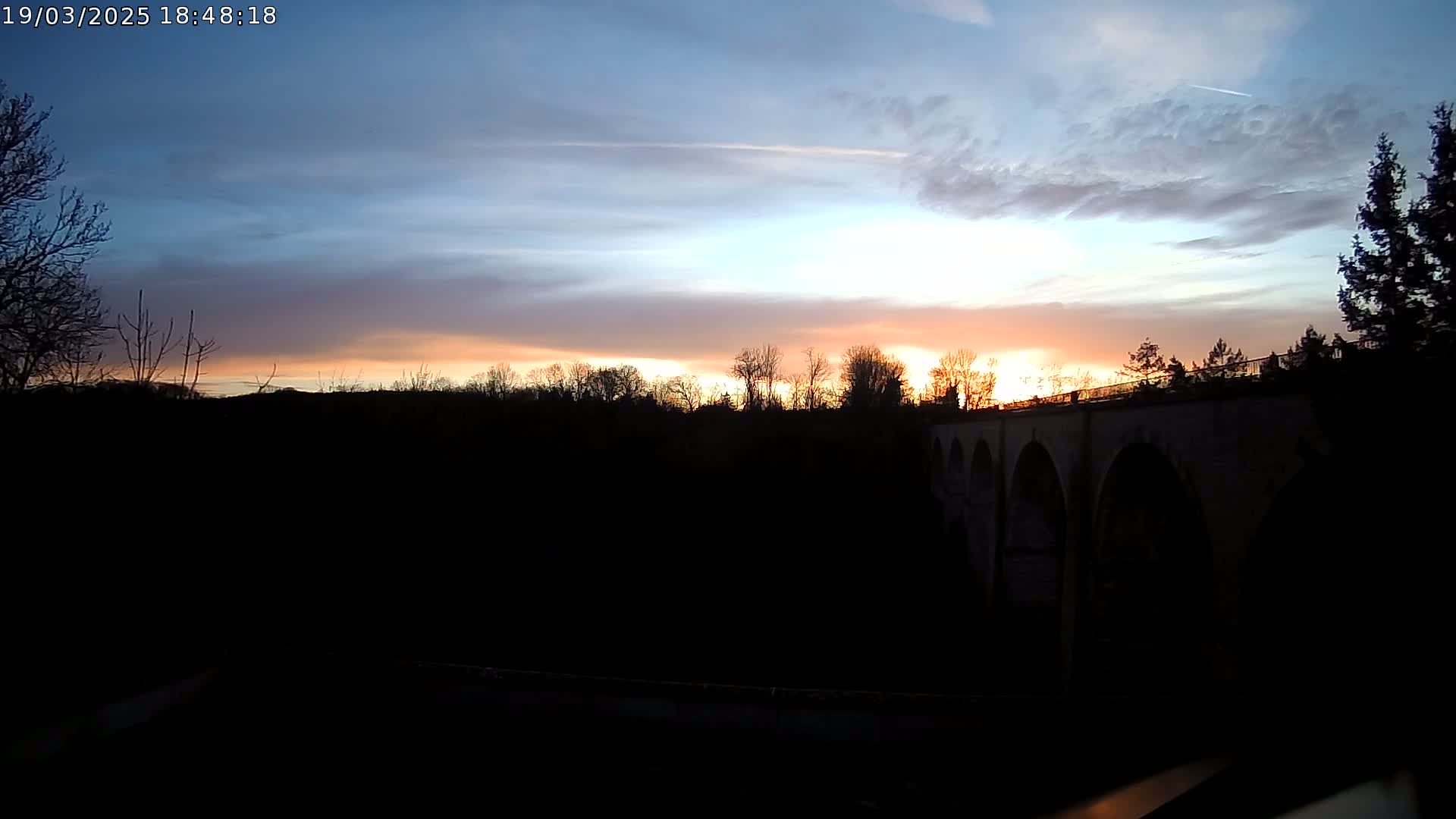 A silhouetted stone bridge is visible against a sunset sky with a mostly clear sky and some scattered clouds.