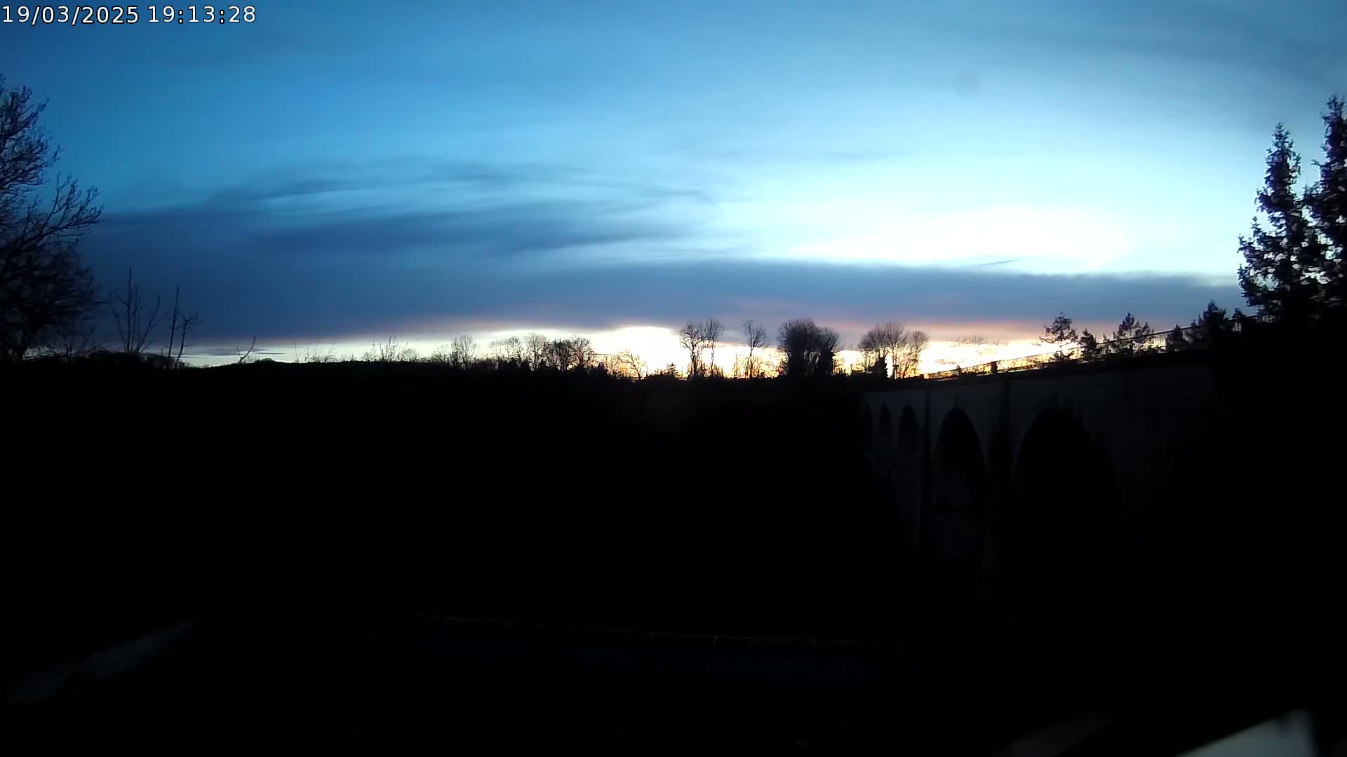 A stone bridge is silhouetted against a twilight sky with thin clouds, as the sun sets behind a line of trees.