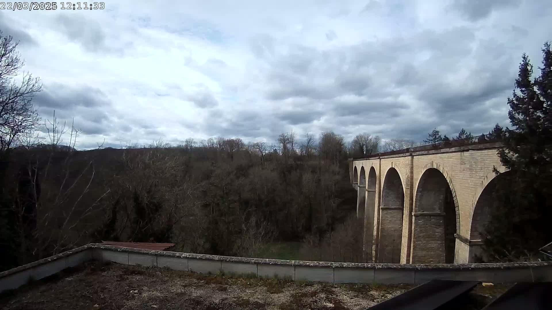 A long stone bridge with multiple arches stretches across a valley of leafless trees under a cloudy sky.