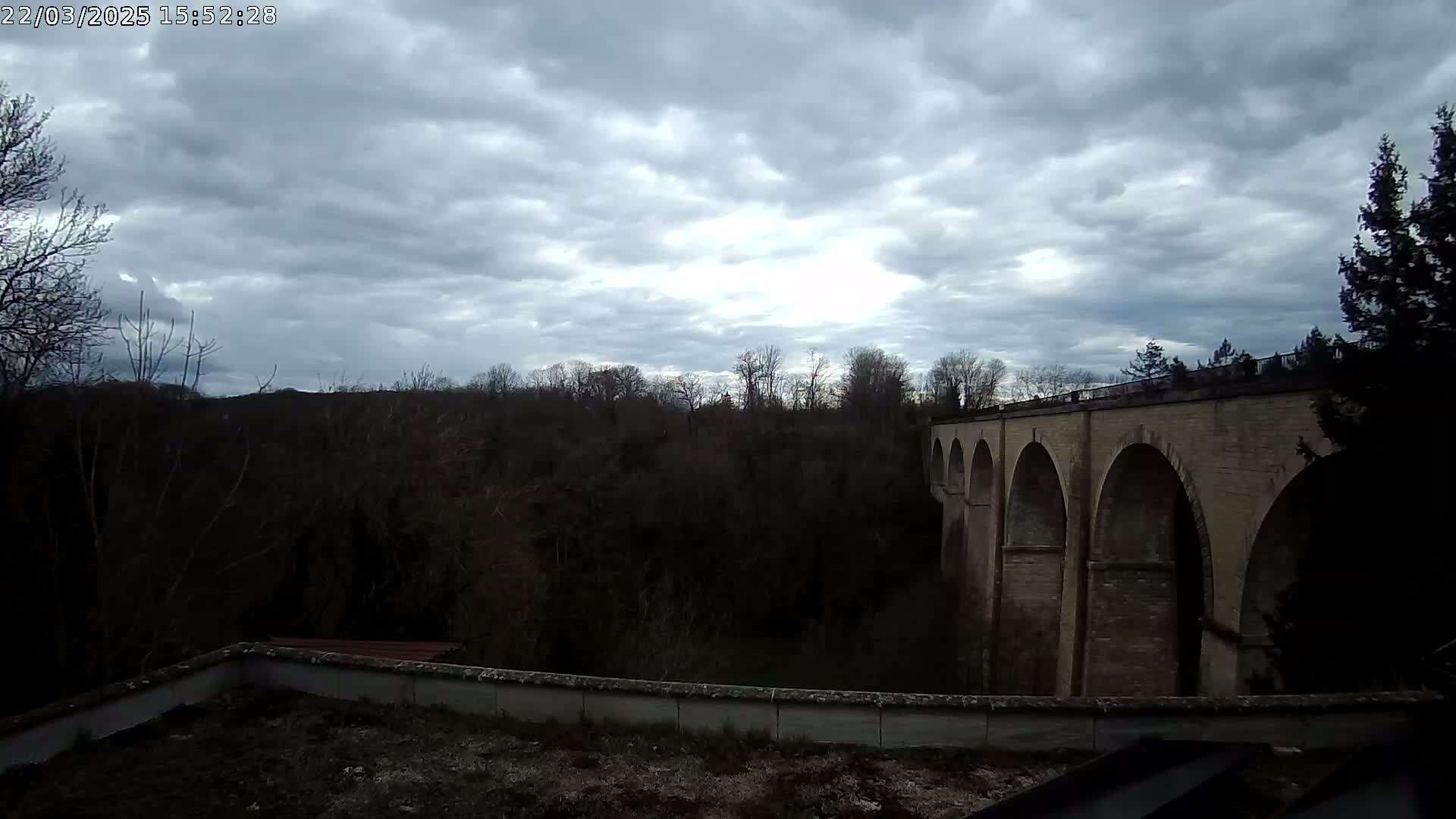 A stone viaduct with multiple arches is seen extending across a wooded valley under a cloudy sky.