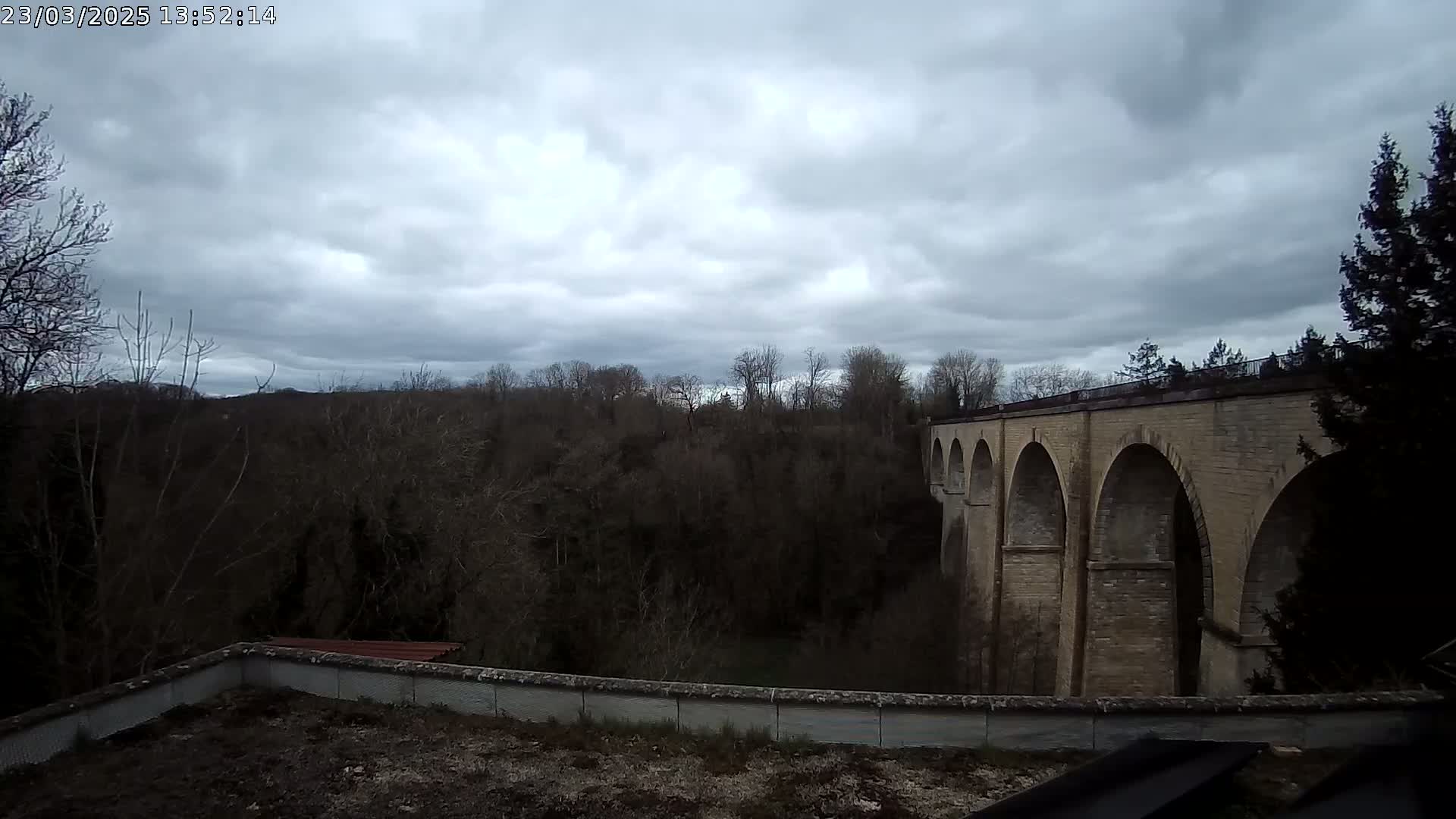 A stone viaduct with multiple arches stands behind a low wall and overlooks a wooded area under a cloudy sky.