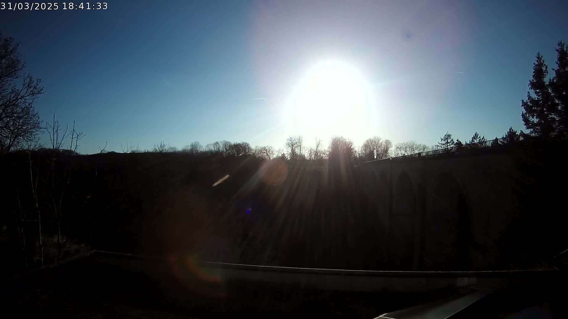 A bright sun shines over a dark, silhouetted stone bridge and trees on a clear day.