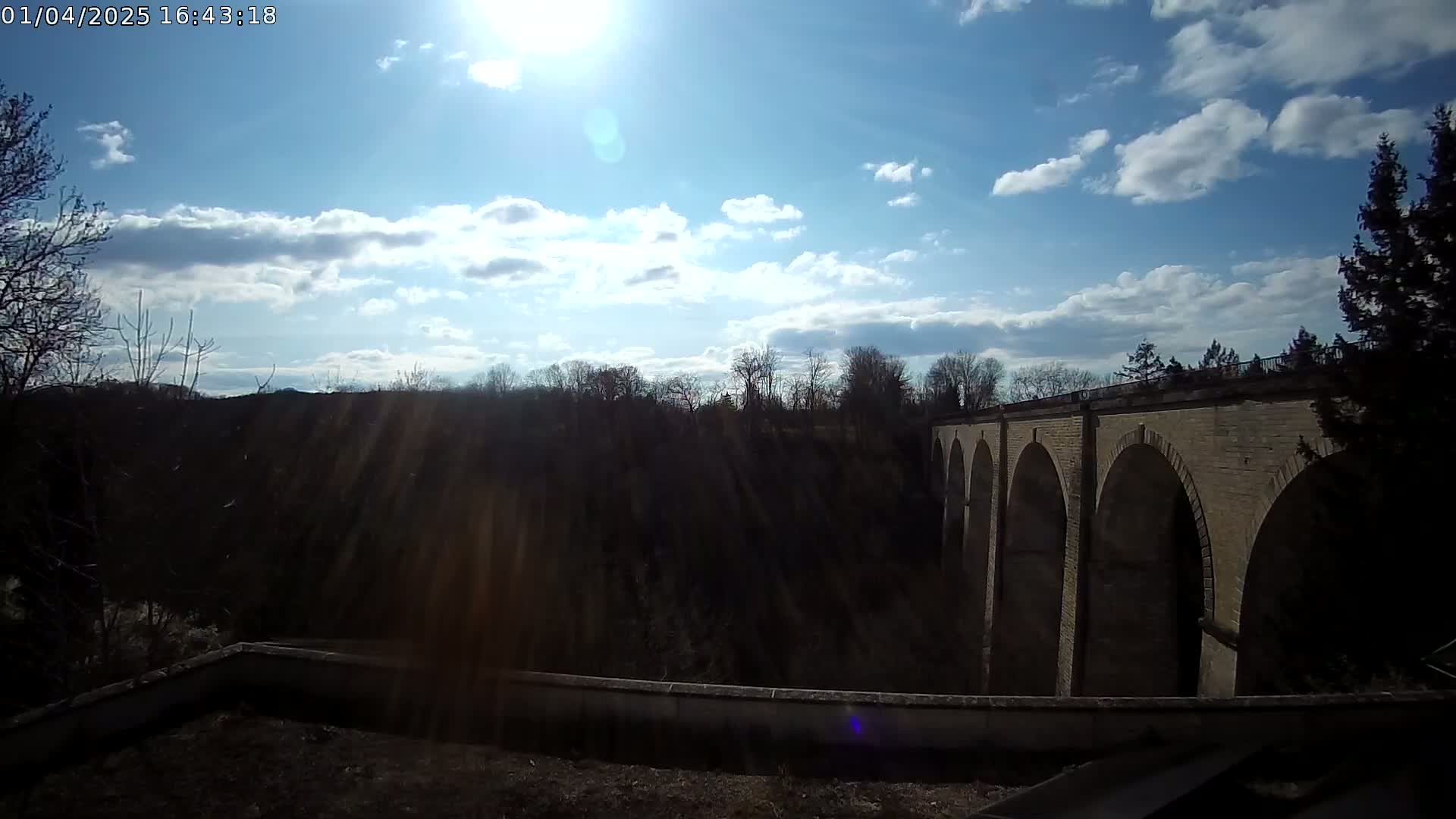 A stone bridge with multiple arches is seen from a distance on a partly sunny day with scattered clouds.