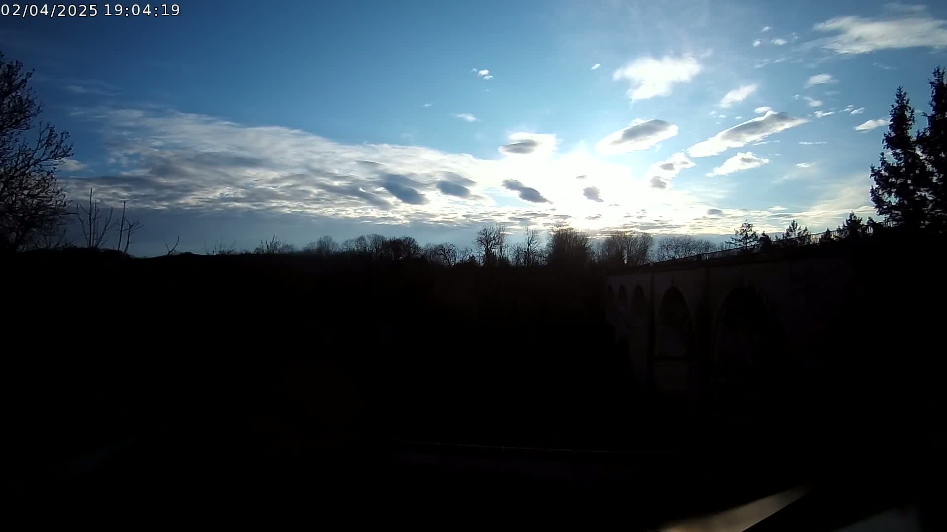 A partly cloudy sky with the sun shining through, behind a silhouetted landscape featuring trees and a bridge.