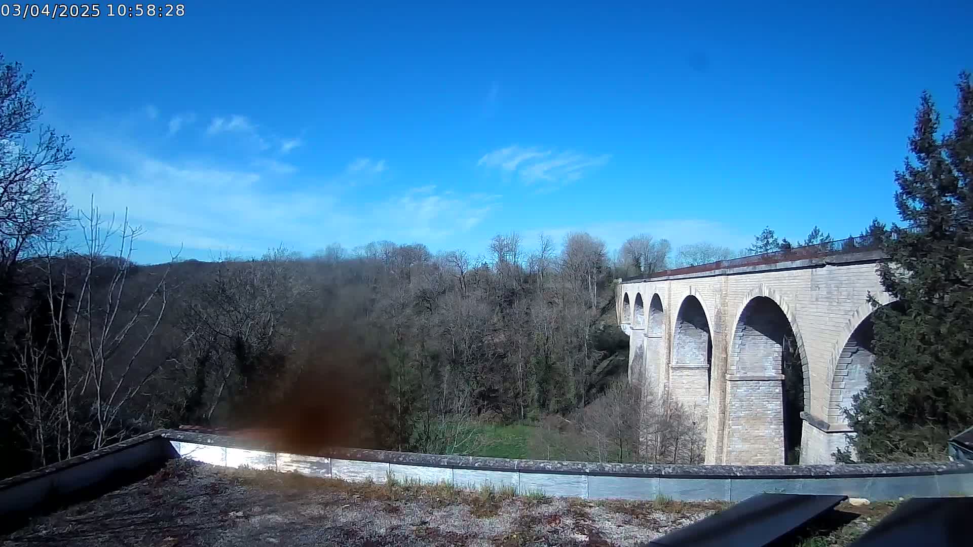 A stone viaduct with multiple arches stretches across a valley filled with leafless trees under a mostly clear blue sky.