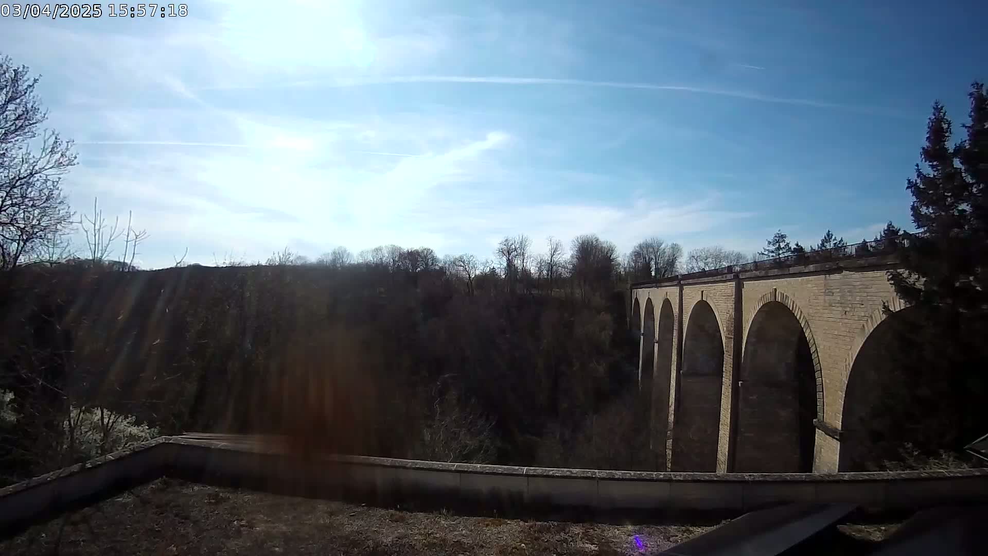 A stone viaduct with multiple arches is seen from a high vantage point on a sunny day with a partly cloudy sky and some trees in the foreground and background.