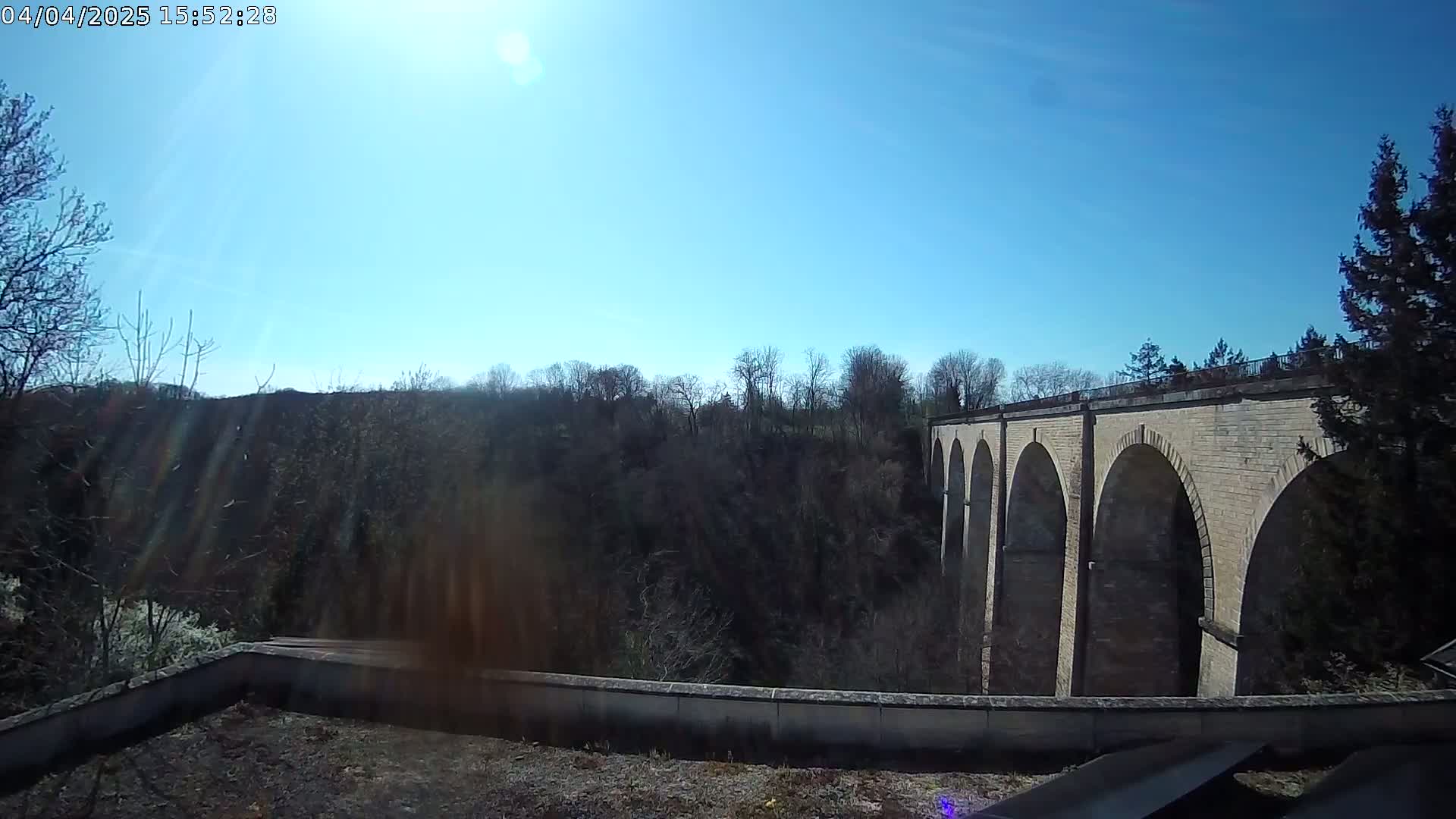 A stone viaduct with multiple arches stretches across a valley of leafless trees under a bright, sunny sky.