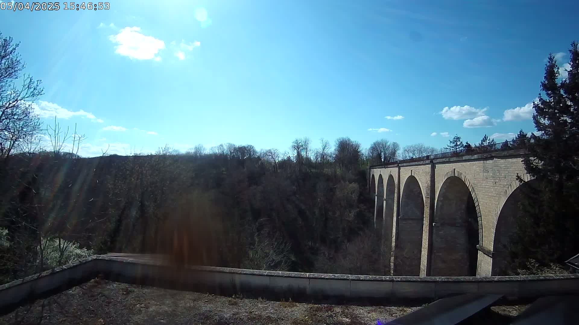 A stone viaduct is seen extending from behind a low wall, under a mostly sunny sky with some scattered clouds, overlooking a wooded valley.