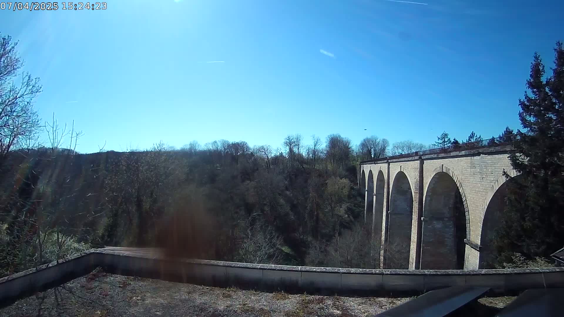 A stone viaduct with multiple arches is visible through a stand of leafless trees under a clear blue sky.