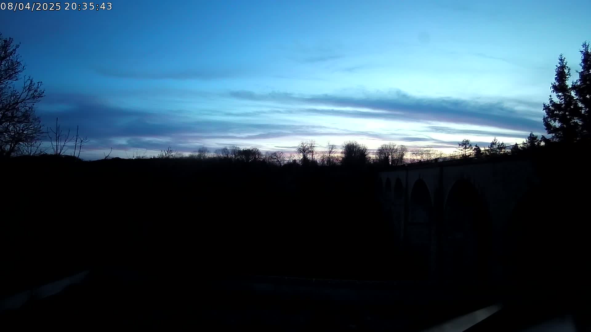 A dark silhouette of trees and a stone arch bridge is visible against a twilight sky with thin, wispy clouds.