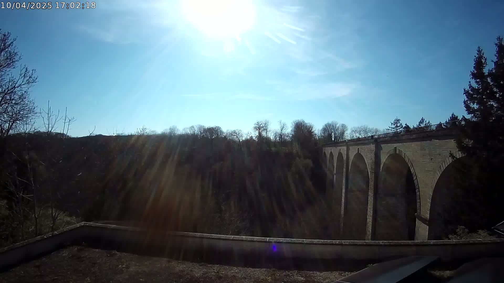 A stone viaduct is visible beyond a line of trees on a sunny day with a mostly clear blue sky.