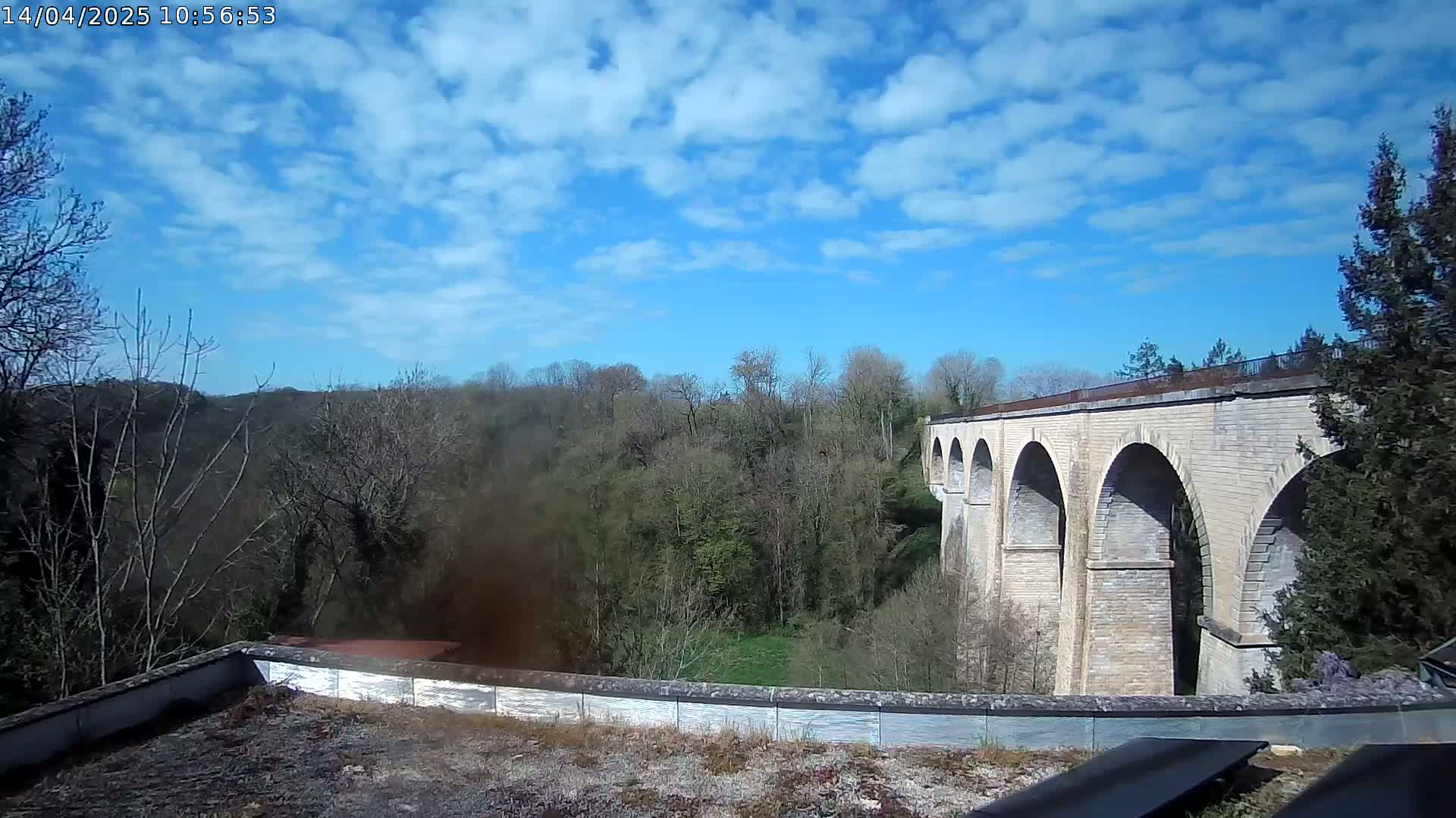 A stone viaduct stretches across a valley of budding trees under a partly cloudy blue sky.