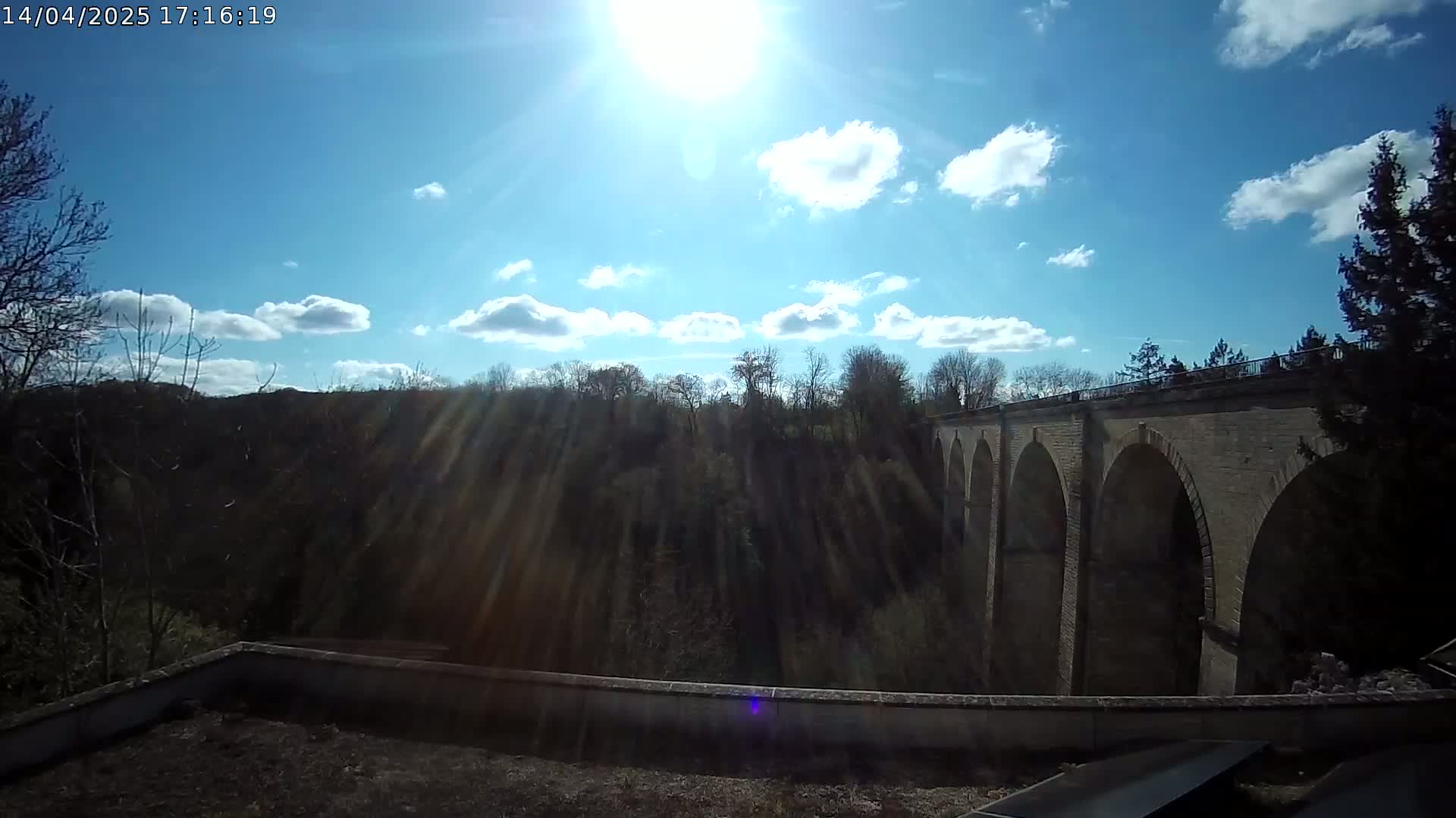 A stone arched bridge spans a wooded valley under a mostly sunny sky with scattered cumulus clouds.