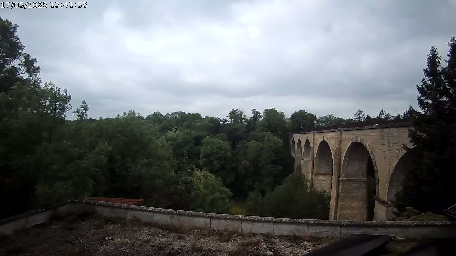 A stone viaduct with multiple arches is partially visible behind a wall and lush green trees under a cloudy sky.