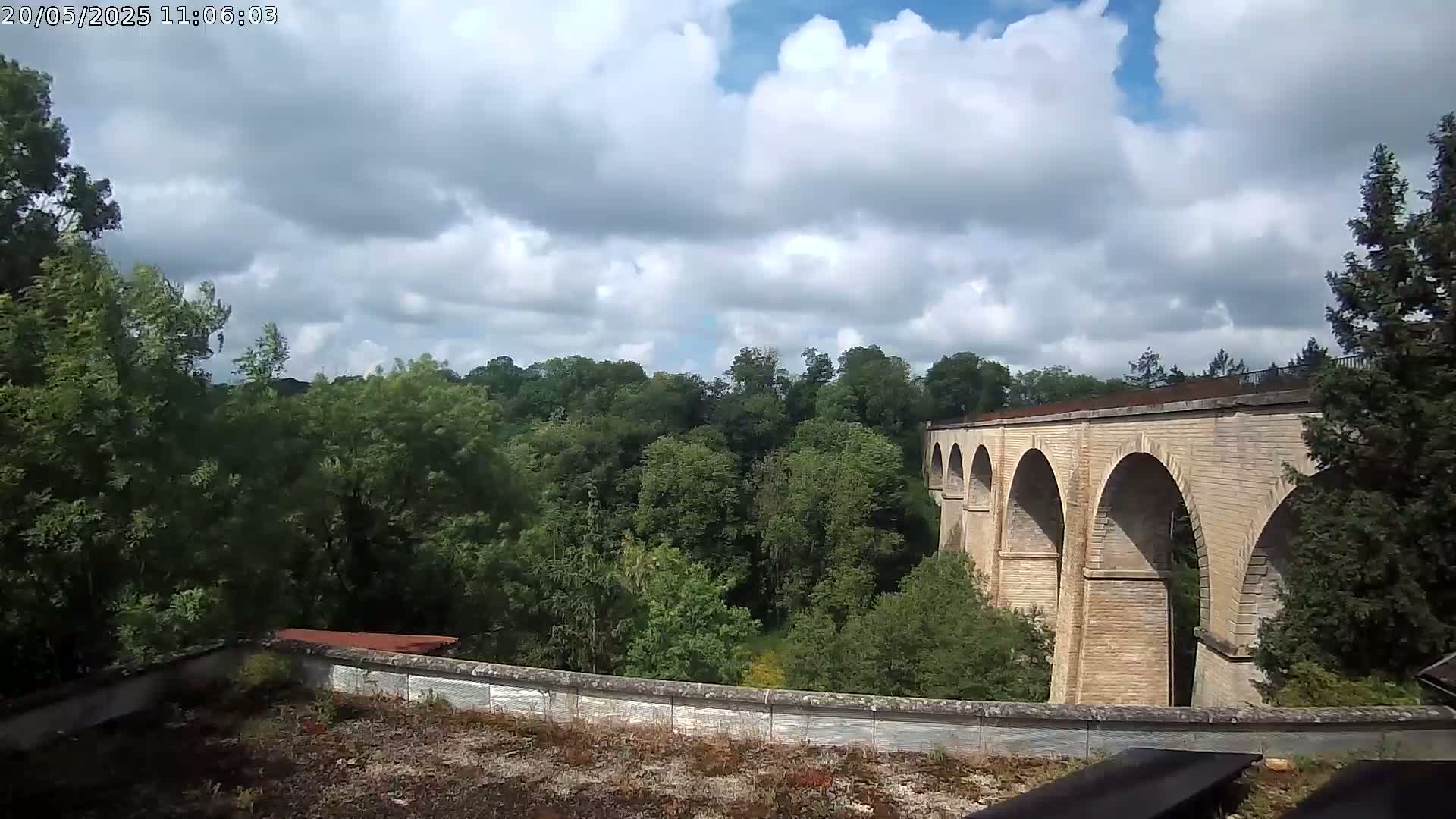A stone viaduct with multiple arches stretches across a verdant valley under a partly cloudy sky.