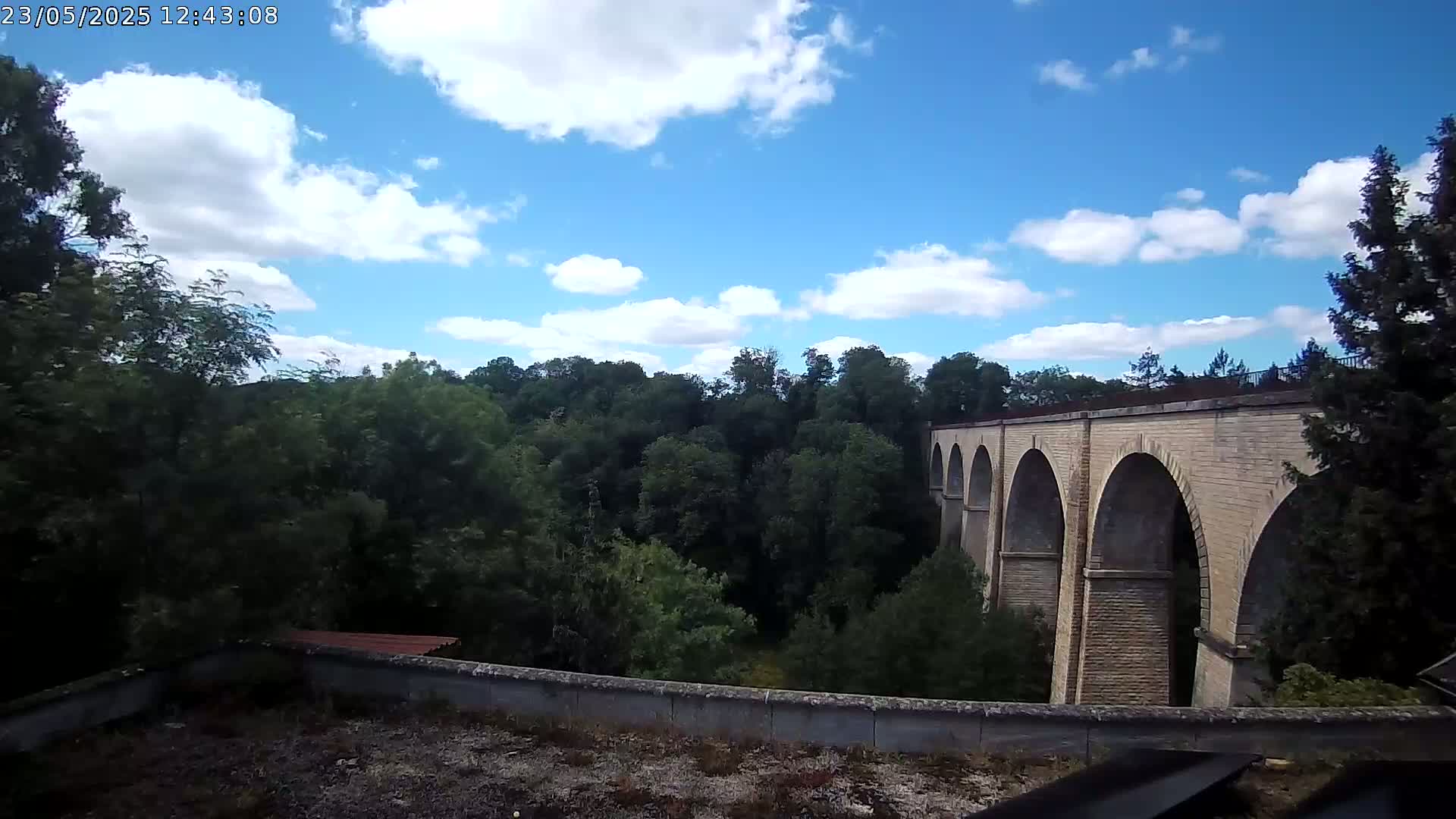 A stone viaduct with multiple arches is visible beyond a line of leafy green trees under a partly cloudy blue sky.