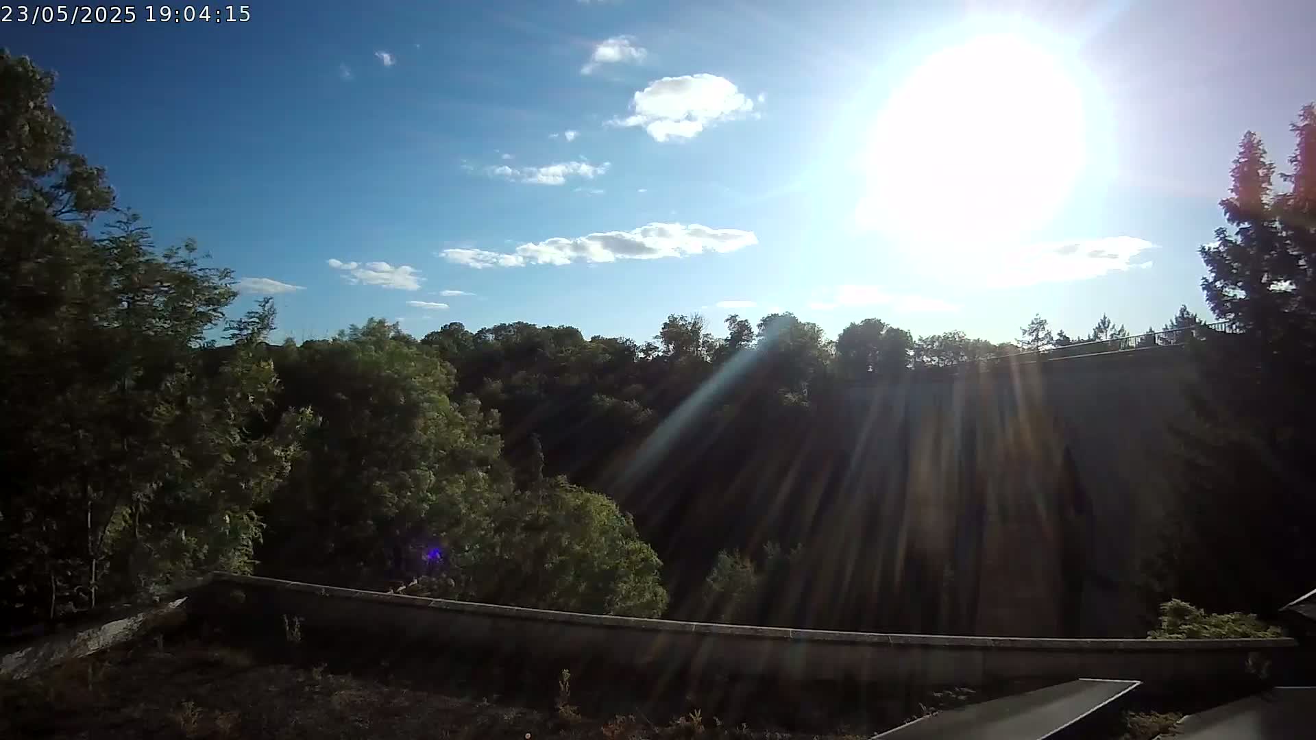 A sunny day with partly cloudy skies is viewed from a high vantage point overlooking a wall and lush green trees.