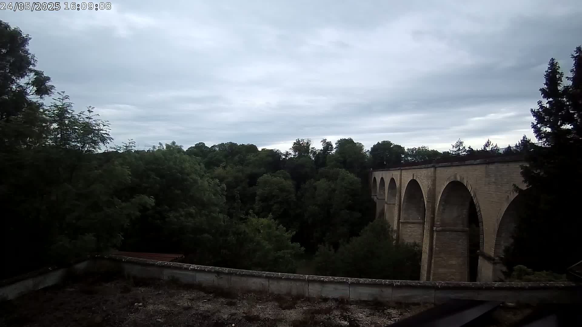 A stone viaduct with multiple arches is visible beyond a line of trees under a cloudy sky.