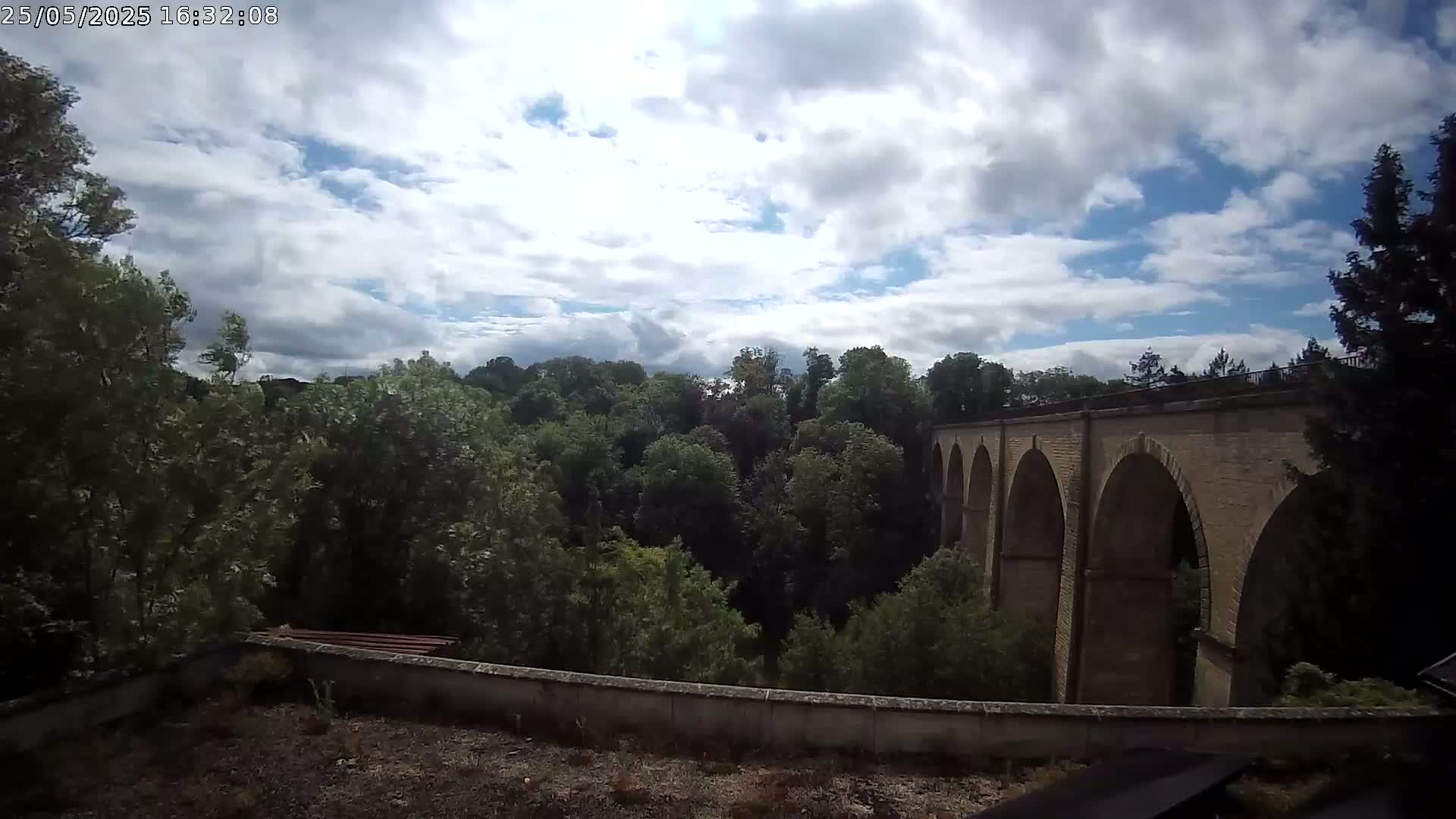 A stone viaduct with multiple arches stretches across a valley filled with lush green trees under a partly cloudy sky.
