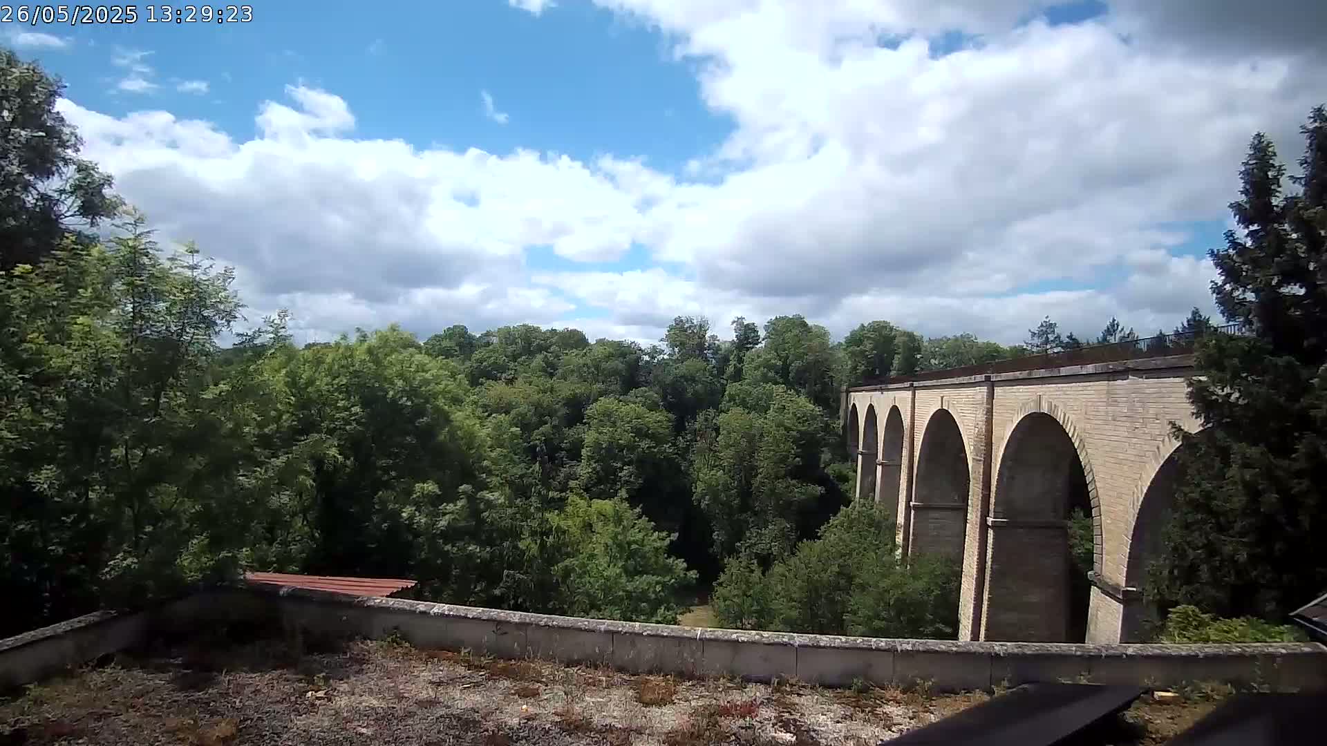A stone viaduct with multiple arches is visible through a lush green forest under a partly cloudy sky.