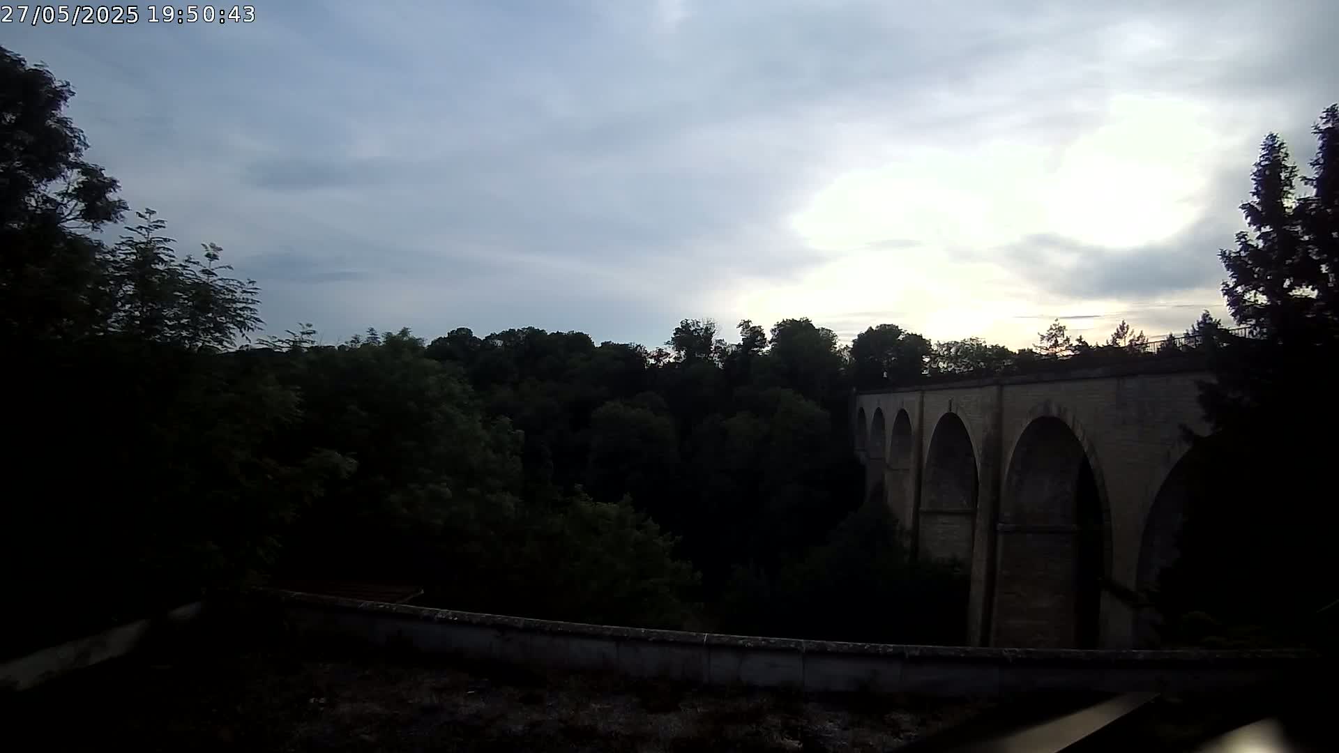 A stone viaduct with multiple arches is visible beyond a line of trees under a partly cloudy sky.