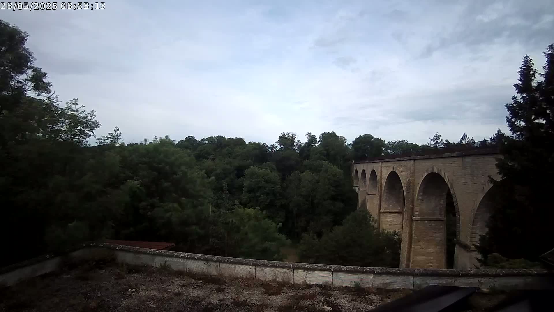 A stone viaduct with multiple arches is visible beyond a wall and a lush green forest under a cloudy sky.