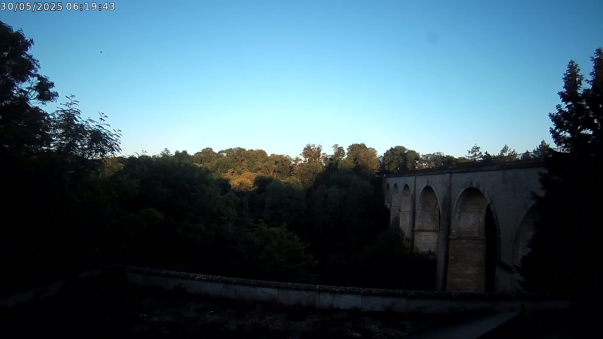 A stone arched bridge is visible through a line of dark green trees under a clear blue sky.
