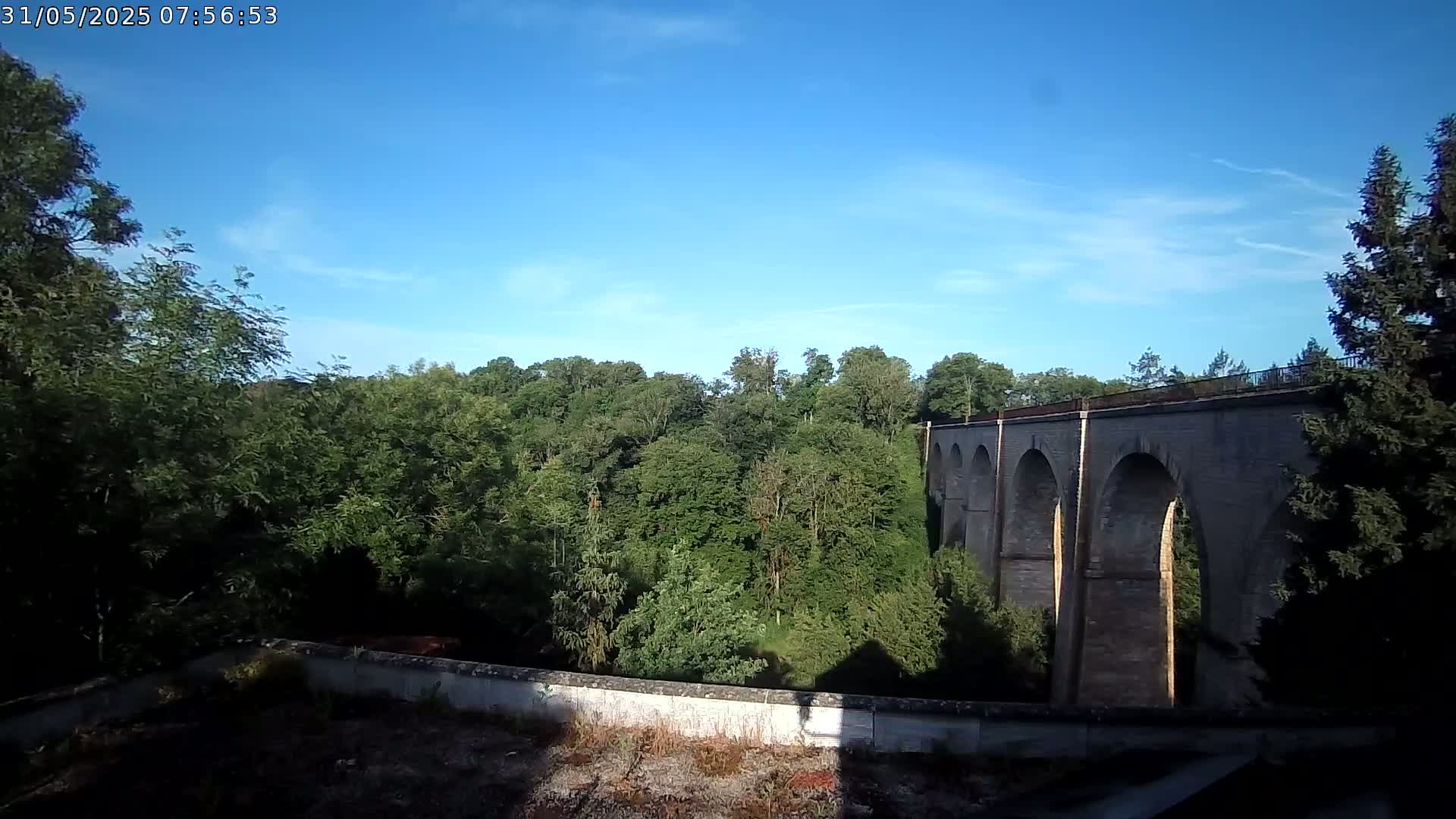 A stone viaduct with multiple arches stretches across a lush green valley under a clear blue sky.