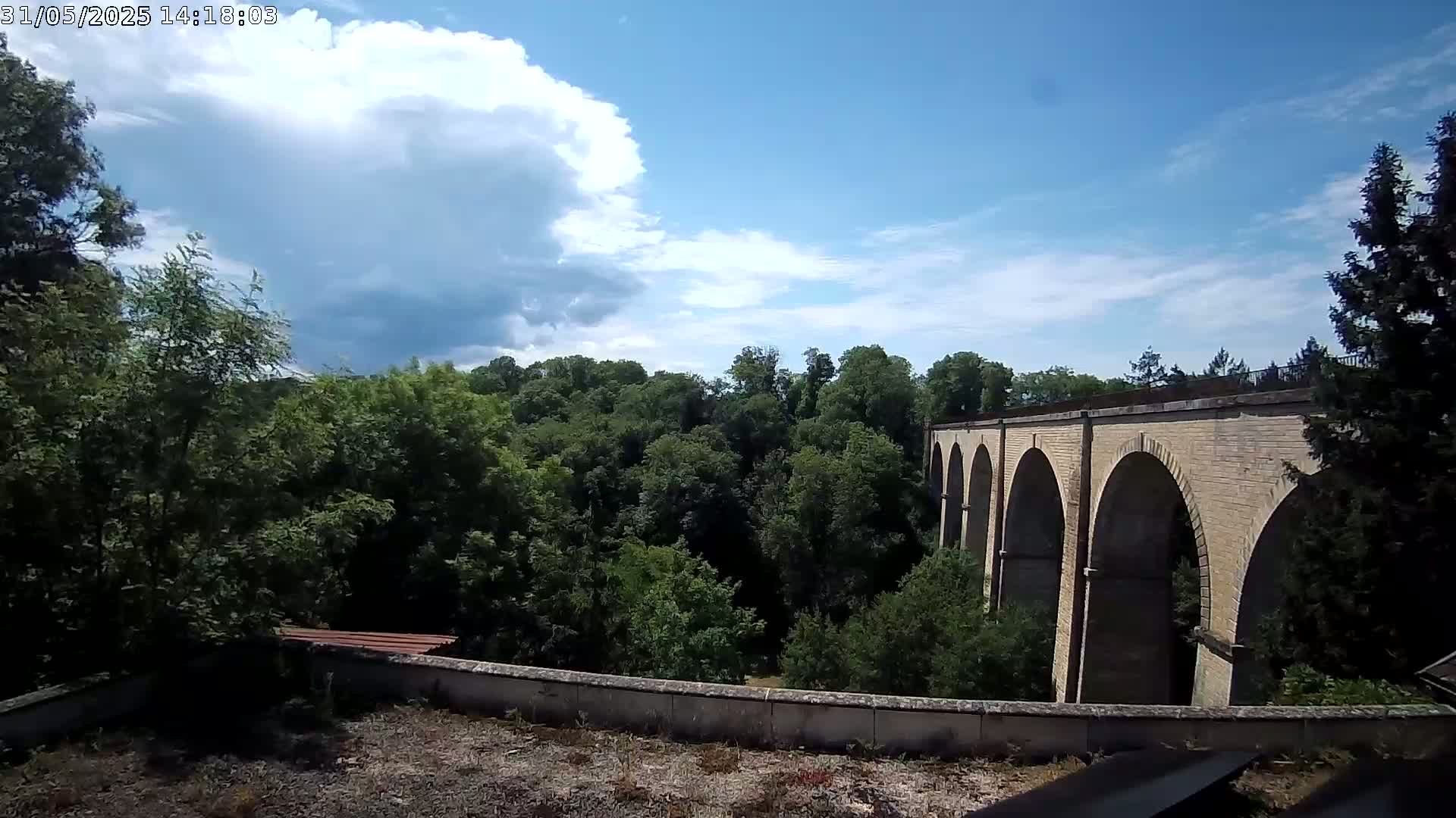 A stone arched viaduct is seen extending from a lush green forest under a partly cloudy blue sky.